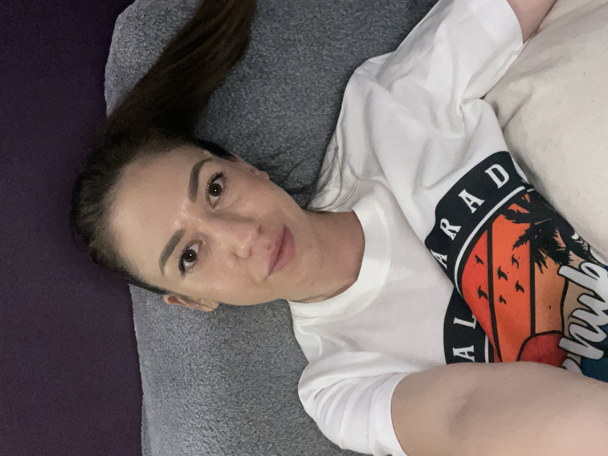 A woman with brown hair lies on a grey surface with a purple edge visible on the left. She wears a white t-shirt with a graphic print on the sleeve. The print features the word "PARADISE" and an image of a sunset with palm trees and birds. She looks directly at the camera with a slight smile. The location is not identifiable as Sofia, Bulgaria from the information given.