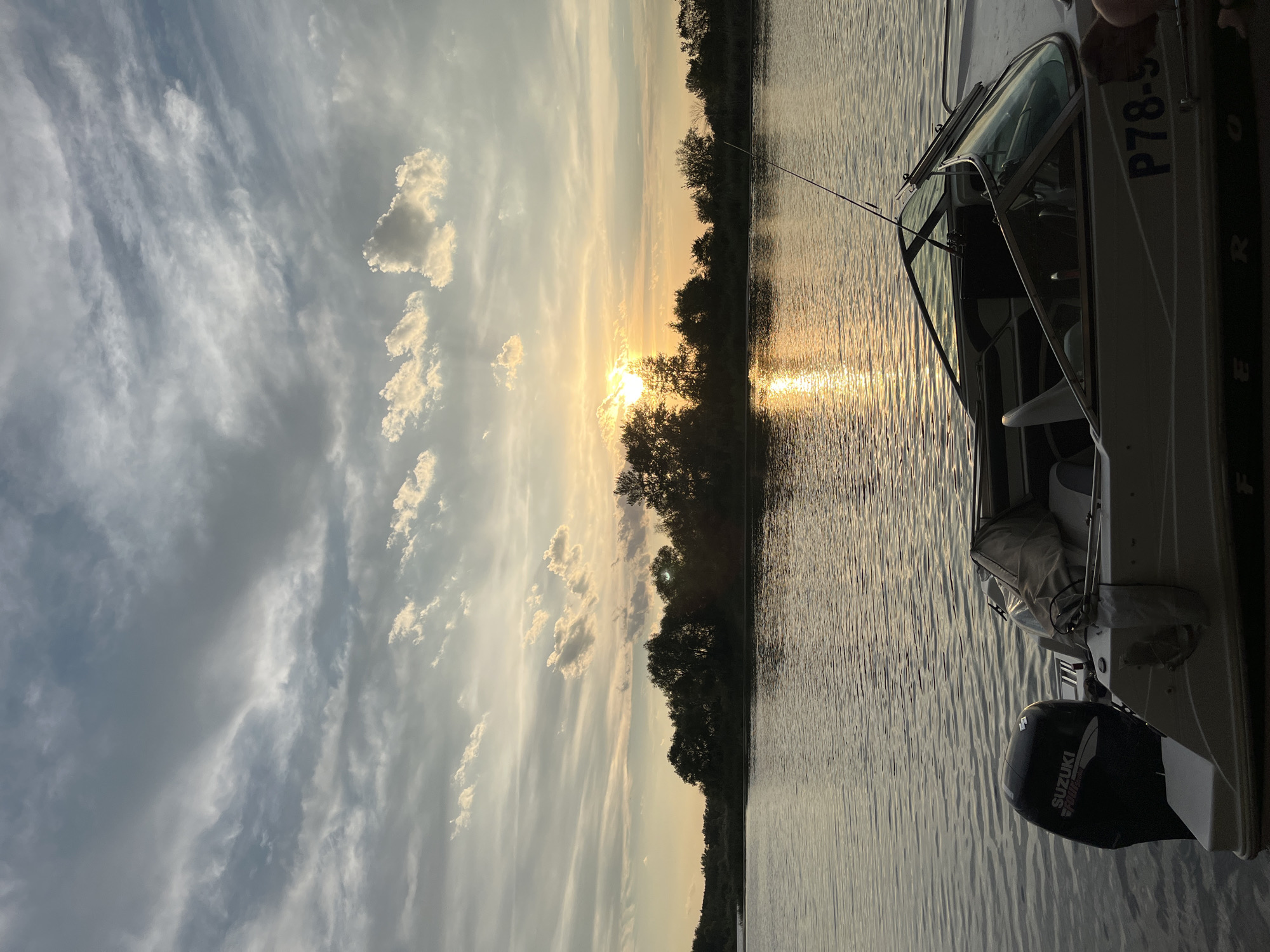 This is a medium shot, taken at eye level, of a motorboat docked on the water at sunset. The boat is white with a dark outboard motor labeled "Suzuki." A fishing rod is visible extending from the back of the boat. The water is calm and reflects the golden light of the setting sun, which is partially obscured by trees on the opposite bank of the river. The sky is filled with scattered clouds, some of which are illuminated by the sun. The scene appears to be set in a natural environment, likely a river or lake, with a wooded shoreline in the background. The location is the city of Shumeyka, Russia.
