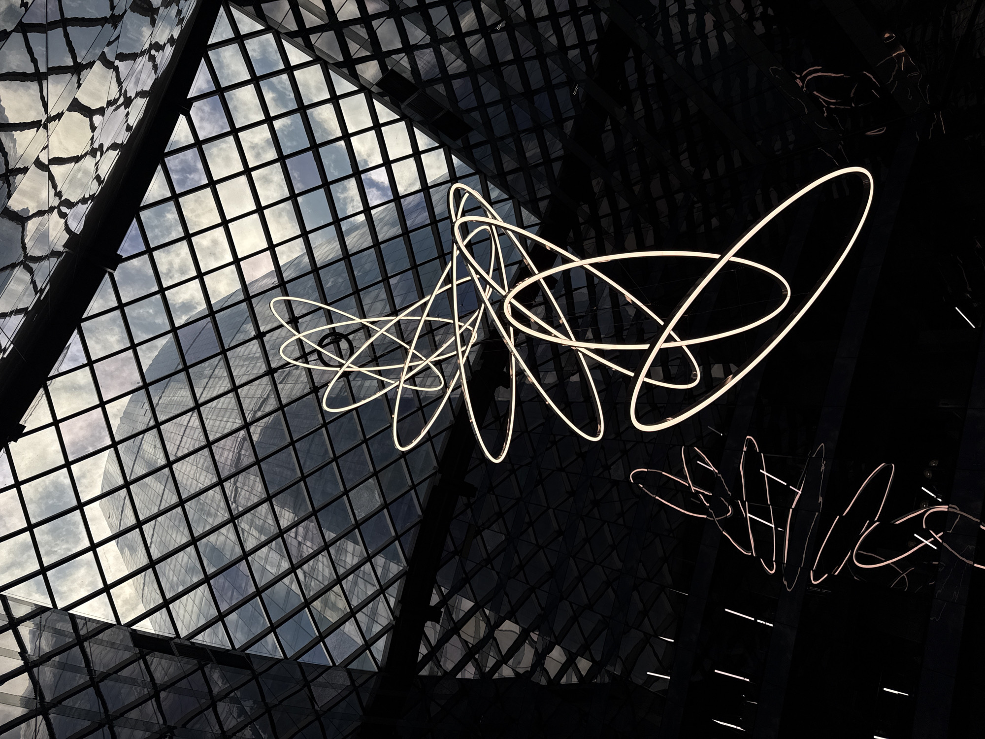 The image depicts an interior view through a complex architectural structure, likely a glass roof or atrium. The left side of the frame reveals a grid pattern of glass panels reflecting a cloudy sky. The right side is significantly darker, suggesting a different lighting condition or a separate part of the building. In the center of the image is a series of interconnected, illuminated light fixtures resembling abstract loops or orbits. Below the illuminated fixture, there is a faint mirrored image of a similar fixture. There are no people visible in the image. There is no information present to connect this image to the city of Khimki, Russia.