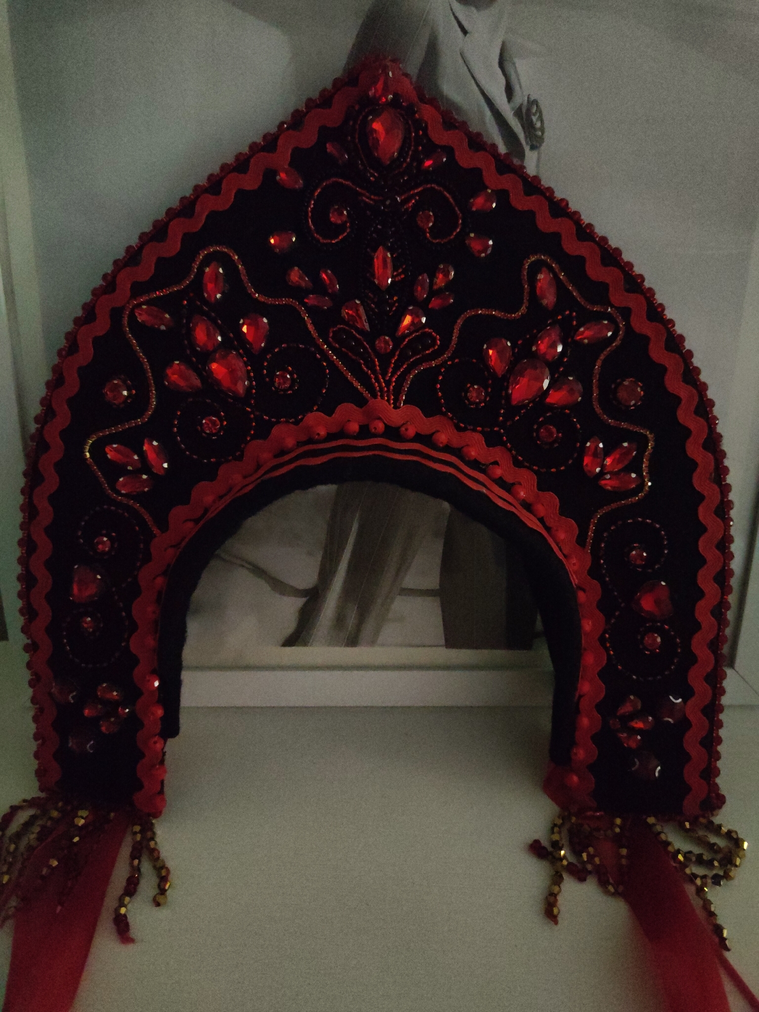 A black fabric kokoshnik, a traditional Russian headdress, is decorated with red jewels and beads. It sits on a white surface in front of a blurred black and white image, likely a photograph or print. The headdress has a scalloped red border and red bead embellishments. There are red fabric tassels with beads hanging from the bottom corners. The location is Unknown, Unknown.