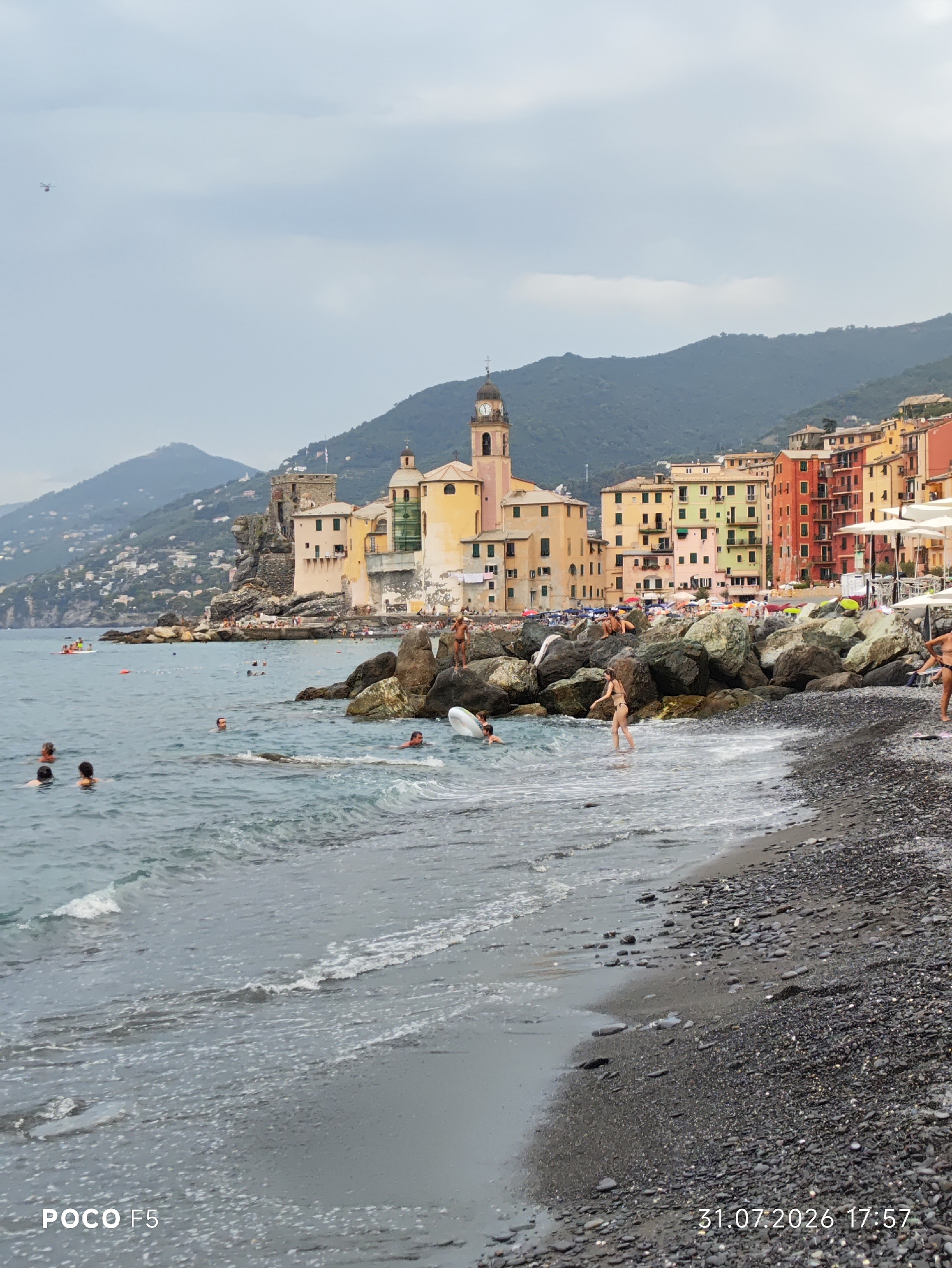 The image shows a view of the beach and town of Camogli, Italy. In the foreground, the choppy blue sea is in motion with small waves breaking on a dark, pebbly beach. Several people are visible in the water, some swimming, others standing. On the right side of the image, a few large rocks are scattered along the shoreline, and a person is standing on one of them. Further back, the beach is lined with beach umbrellas and lounge chairs. Behind the beach, colorful buildings rise up the hillside, with a prominent church tower featuring a clock. Mountains form the backdrop to the town, and the sky is overcast. A small red helicopter is visible in the sky far in the distance. The bottom of the image has text overlays indicating "POCO F5" and "31.07.2026 17:57".