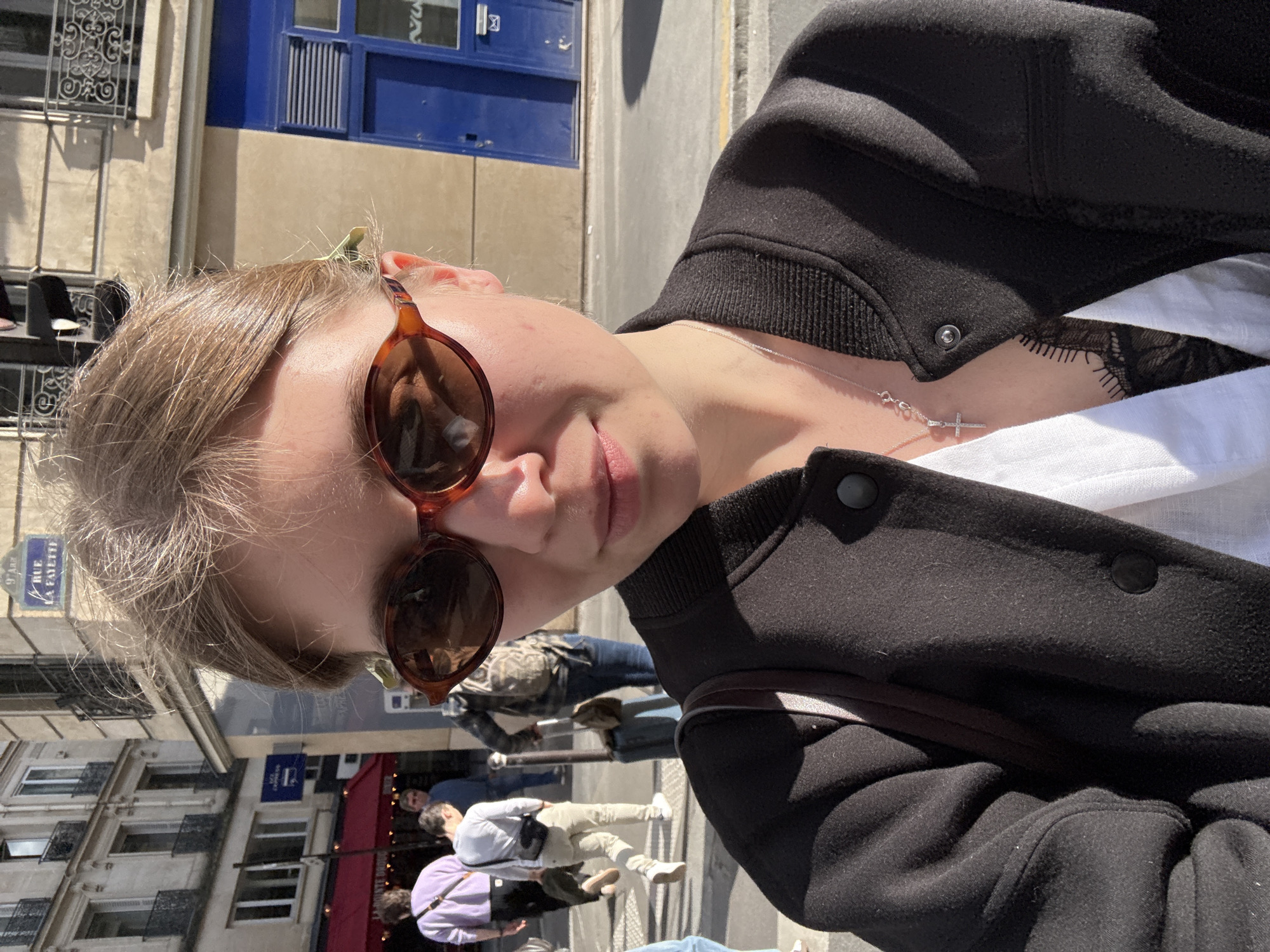 A close-up, tilted selfie captures a young woman outdoors on a sunny day in Paris, France. She is wearing brown-framed, dark-lensed sunglasses, a black bomber-style jacket with ribbed cuffs, and a white collared shirt revealing a hint of black lace underneath. A silver chain necklace with a small cross pendant hangs around her neck. Her light brown hair is styled away from her face, with some lighter strands visible.
The background shows a bustling urban street with several blurred pedestrians walking past buildings typical of Parisian architecture. One prominent blue street sign, partially obscured, clearly displays "RUE LA FAYETTE" and "9 ARR", confirming the location as the 9th arrondissement of Paris. The bright sunlight and distinct shadows indicate it is daytime. Other background elements include buildings with ornate window railings, a blue facade on a building, and hints of red awnings from establishments. The woman's expression appears relaxed, with her lips slightly parted.
