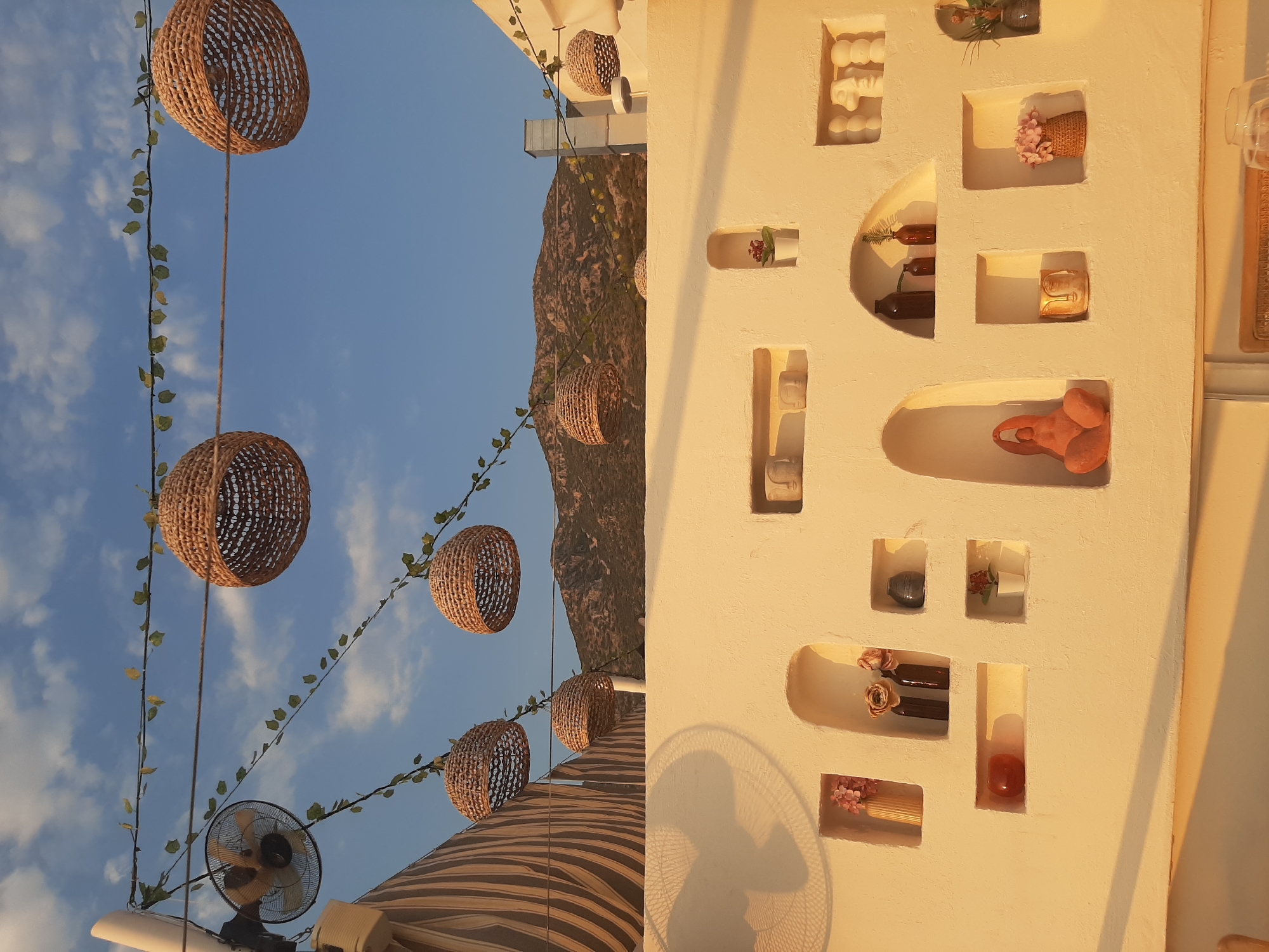 The image depicts an outdoor patio area, possibly a cafe or restaurant, in Kaş, Turkey. The setting is characterized by a white, textured wall with several recessed niches displaying decorative items. These items include various vases, a small potted plant, a terracotta figurine, and decorative facial sculptures. Above the wall, a clear blue sky with scattered clouds serves as the backdrop. Hanging from wires strung across the space are several woven, basket-like pendant lights. A fan is visible mounted on a pole on the left side of the image, and a striped awning is also partially visible. In the distance, hills and mountains form the horizon. The lighting suggests it is either early morning or late afternoon, casting warm, golden light and shadows. No people are visible in the frame.