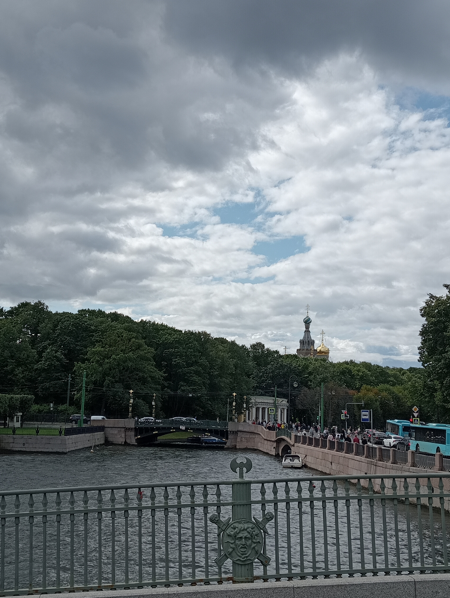The image shows a view of the city of Saint Petersburg, Russia, likely taken from a bridge overlooking a canal. In the background, a church with golden domes is visible, suggesting it is a prominent landmark. The sky is overcast with dramatic clouds.
On the canal, there are several boats, including a light-colored smaller vessel and a larger blue bus-like boat, possibly a tourist boat. The water is choppy, indicating wind or current.
A bridge spans the canal, with people walking along its pedestrian walkway. Cars are seen on the roadway of the bridge, and there are traffic signals. The bridge itself appears to be made of stone with a decorative railing in the foreground.
Dense green trees line the banks of the canal and the city skyline. The overall atmosphere suggests a somewhat gray but active day in the city. The presence of the church with golden domes strongly points to the location being near a significant religious site, potentially the Church of the Savior on Spilled Blood, given the characteristic domes and the canal setting.