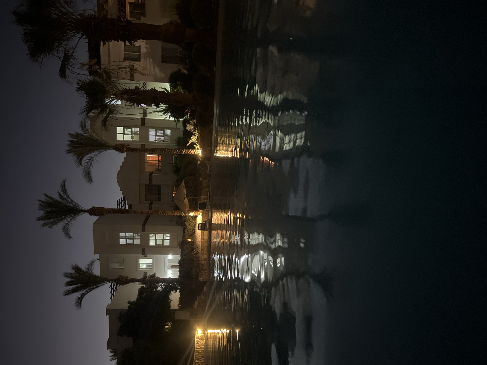 A nighttime exterior shot captures a swimming pool in the foreground, with its surface reflecting the lights of a resort complex situated in Sharm El Sheikh, Egypt. The resort features modern, multi-story buildings with illuminated windows, surrounded by lush greenery and tall palm trees that are characteristic of the region. The water in the pool is dark, with ripples distorting the reflections of the buildings and palm trees, creating an abstract pattern of light and shadow. The overall atmosphere is serene and tranquil, with the artificial lighting creating a warm glow against the dark sky. The scene depicts a typical resort setting in a warm climate, designed for relaxation and leisure.