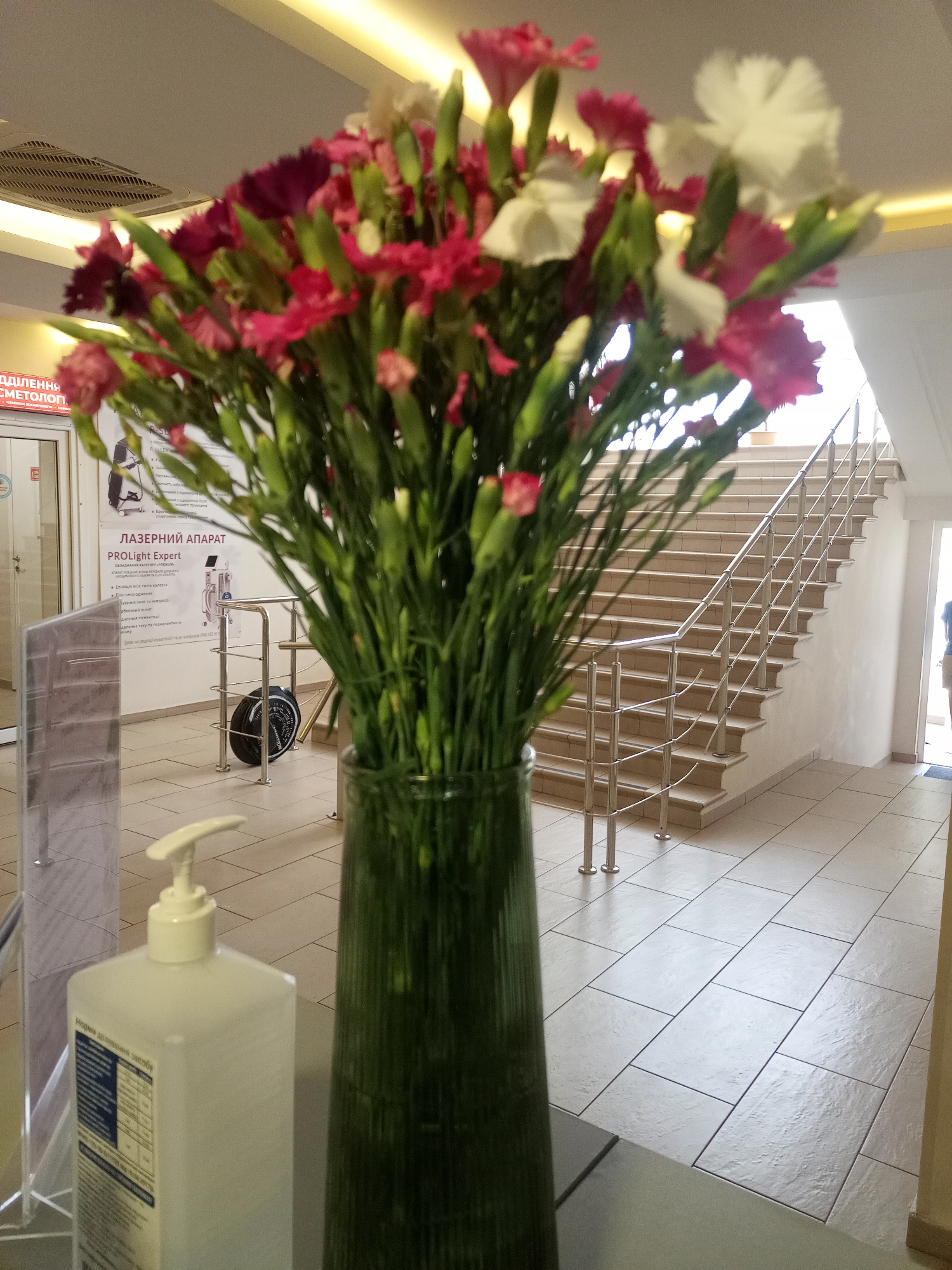 The image shows a bouquet of pink and white carnations in a tall green glass vase, placed on a counter or table in what appears to be a public building lobby. To the left of the flowers, a white dispenser of liquid soap or hand sanitizer is visible. In the background, a staircase with a metal railing leads upwards, and a poster or advertisement is mounted on the wall next to the stairs. The floor is tiled. The lighting is bright, suggesting an indoor environment with artificial lights. The scene is static, with no discernible activity or people in focus.