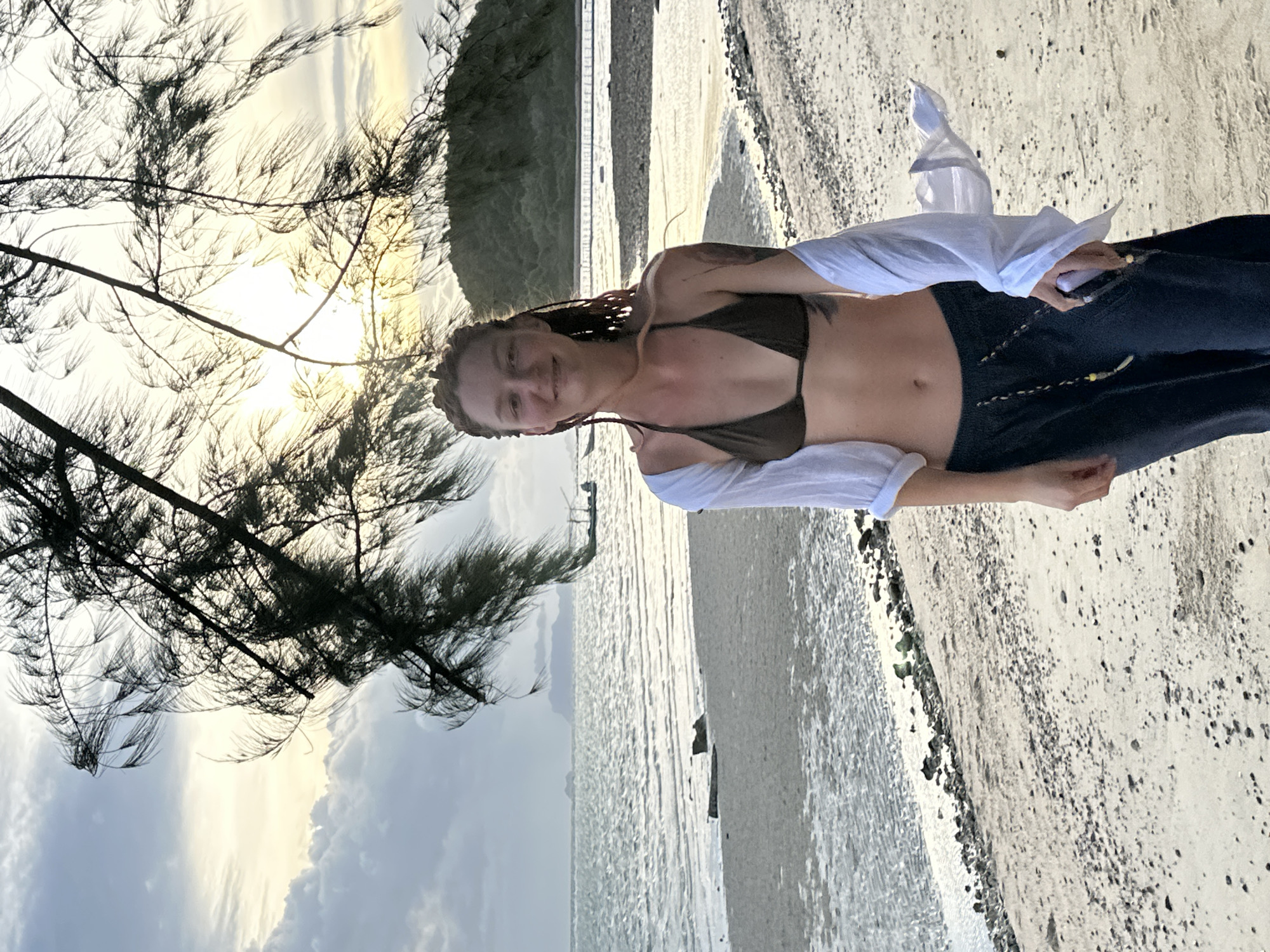 A young woman with braided hair smiles at the camera as she stands on a sandy beach. She is wearing a dark bikini top and a loose white shirt. Her dark blue jeans are unbuttoned, revealing her midriff. A tattoo is visible on her upper arm. The beach is composed of light-colored sand with small pebbles and rocks scattered across it. The ocean stretches out behind her, with calm, shimmering water. In the distance, a lush green hillside rises from the water.
To the left of the woman, a dark pine tree with long, wispy needles extends into the frame, its branches silhouetted against a bright, hazy sky. The sun is visible through the tree, creating a warm glow that suggests it is either early morning or late afternoon. The sky is a mix of pale yellow, white, and soft grey clouds, indicating possibly overcast or hazy conditions. The overall scene is peaceful and serene. The location context places the image in the Ko Samui District of Thailand. There is no visible text in the image.
