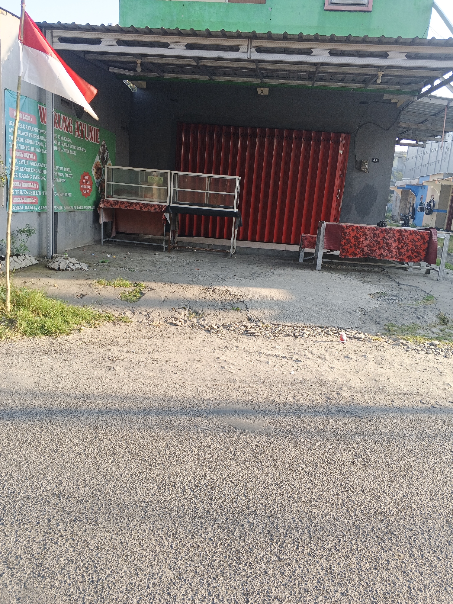 The image shows the exterior of a small shop or warung in Krian, Indonesia, with a red corrugated metal roll-up door in the background. To the left, a flagpole displays the Indonesian flag, red over white. A green sign with white lettering is visible, reading "WRUNG AYUNE" in large font, with smaller text listing food and drink items. In front of the warung, there are two display tables with glass tops, and another table covered with a red floral patterned cloth. The ground in front of the warung is a mix of dirt and gravel, with some patches of grass. The warung appears to be situated along a road, as evidenced by the asphalt in the foreground. The sky is bright, indicating daylight. No people are visible in the scene.