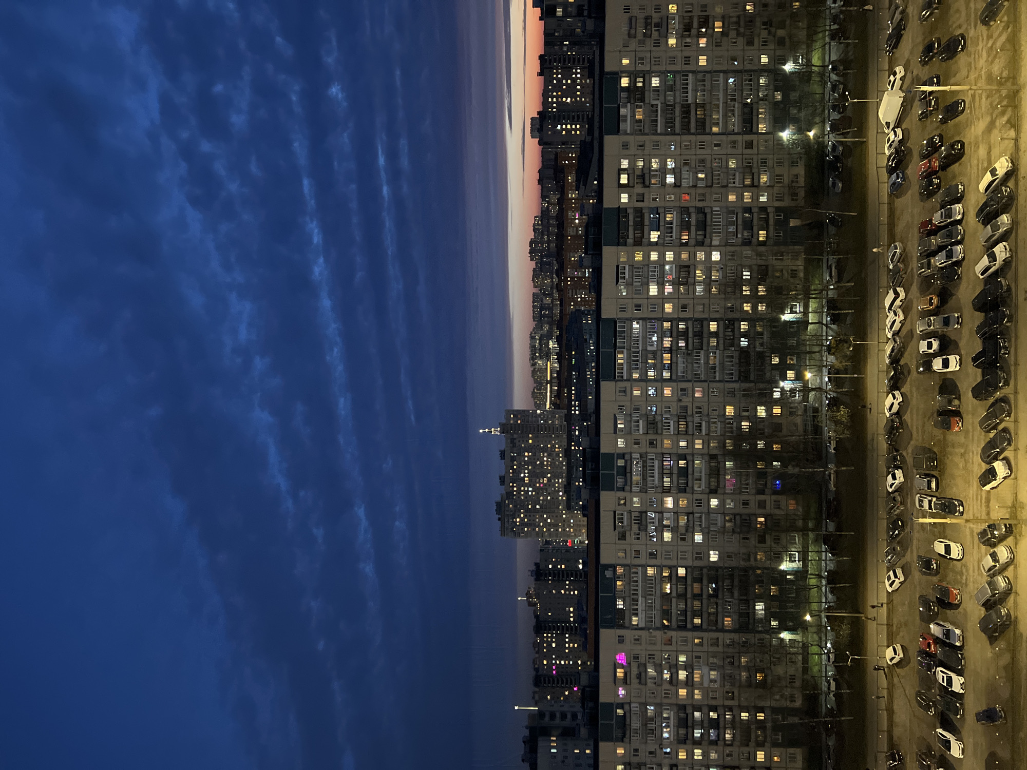 An eye-level, medium shot of a city landscape at dusk or night. The horizon line extends horizontally through the center of the frame, separating the sky from the urban landscape.
On the left side of the image, the sky is deep blue with visible cloud patterns. The right side of the image features a dense collection of apartment buildings, likely in Saint Petersburg, Russia. Many windows are illuminated, suggesting that it is either early morning or late evening.
Adjacent to the buildings on the far right is a parking lot filled with cars. Streetlights are visible, casting light onto the vehicles and the surrounding area.