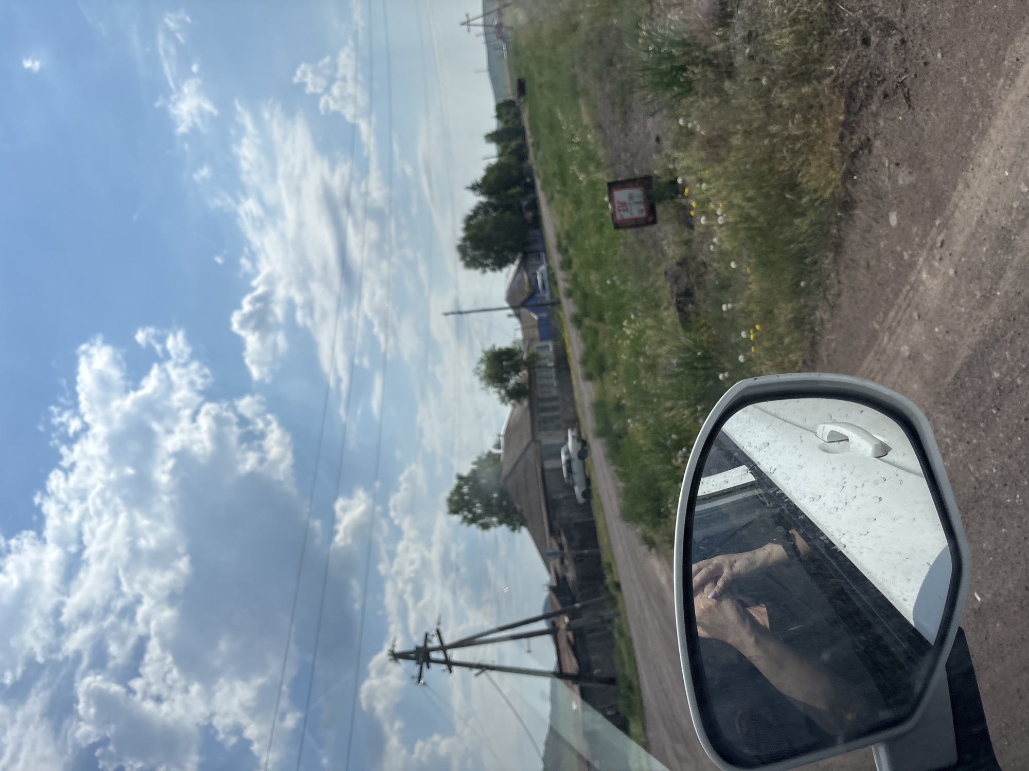 The image shows a rural scene from the perspective of a car passenger, as indicated by the side-view mirror reflecting the passenger's hands. The setting appears to be a village with a dirt road. In the background, there are several houses with low-pitched roofs, some with wooden fences. A power line tower with multiple wires stands prominently on the left. A white car is parked in front of one of the houses. The landscape is characterized by sparse vegetation, with grass and some wildflowers along the roadside. The sky is blue with scattered white clouds, suggesting fair weather. A red and white sign with some markings is visible on the right side of the road, near a small patch of yellow wildflowers. This scene is consistent with a rural location such as Троицкое, Russia, likely a village outside a larger urban center.