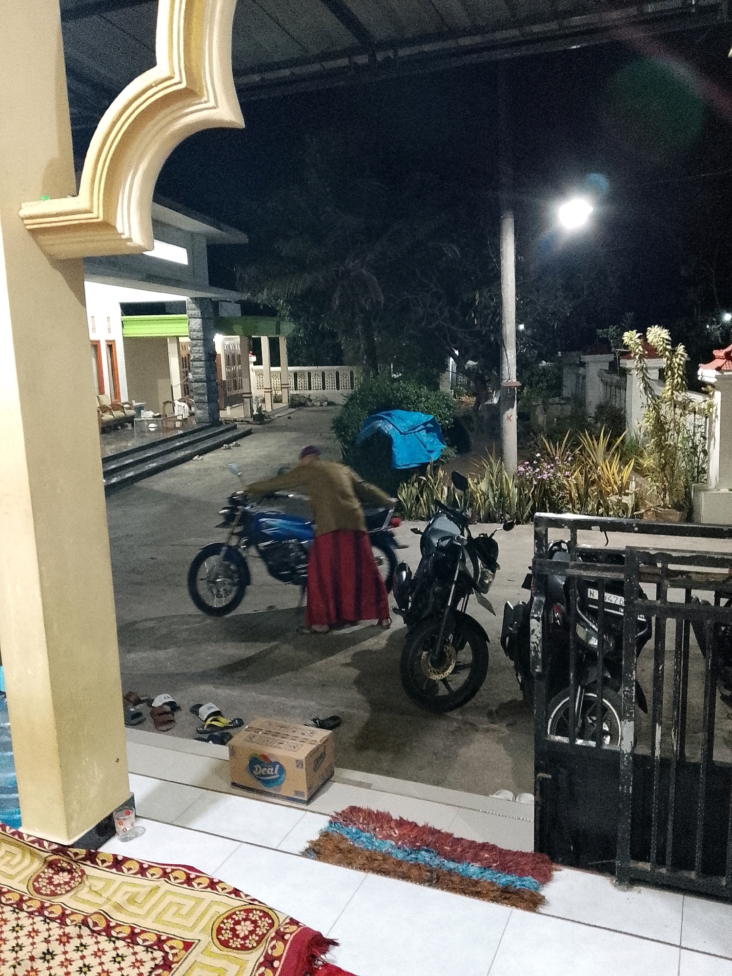 The image depicts an outdoor scene at night, likely a residential area in Rejoyoso, Indonesia. A person wearing a light brown top and a long red skirt is standing next to a blue motorcycle. The motorcycle appears to be older model, with a visible exhaust pipe and spoked wheels. In the background, a building with a green awning and a white decorative facade is visible. There are several other motorcycles parked in the scene, one of which is partially obscured by a black metal gate. Various items are scattered on the ground, including a cardboard box labeled "Deal," several pairs of sandals, and a colorful rug placed on the tiled floor in the foreground. A street light illuminates the scene from the upper right. The overall atmosphere suggests a casual, nighttime setting, possibly a driveway or an area outside a home.