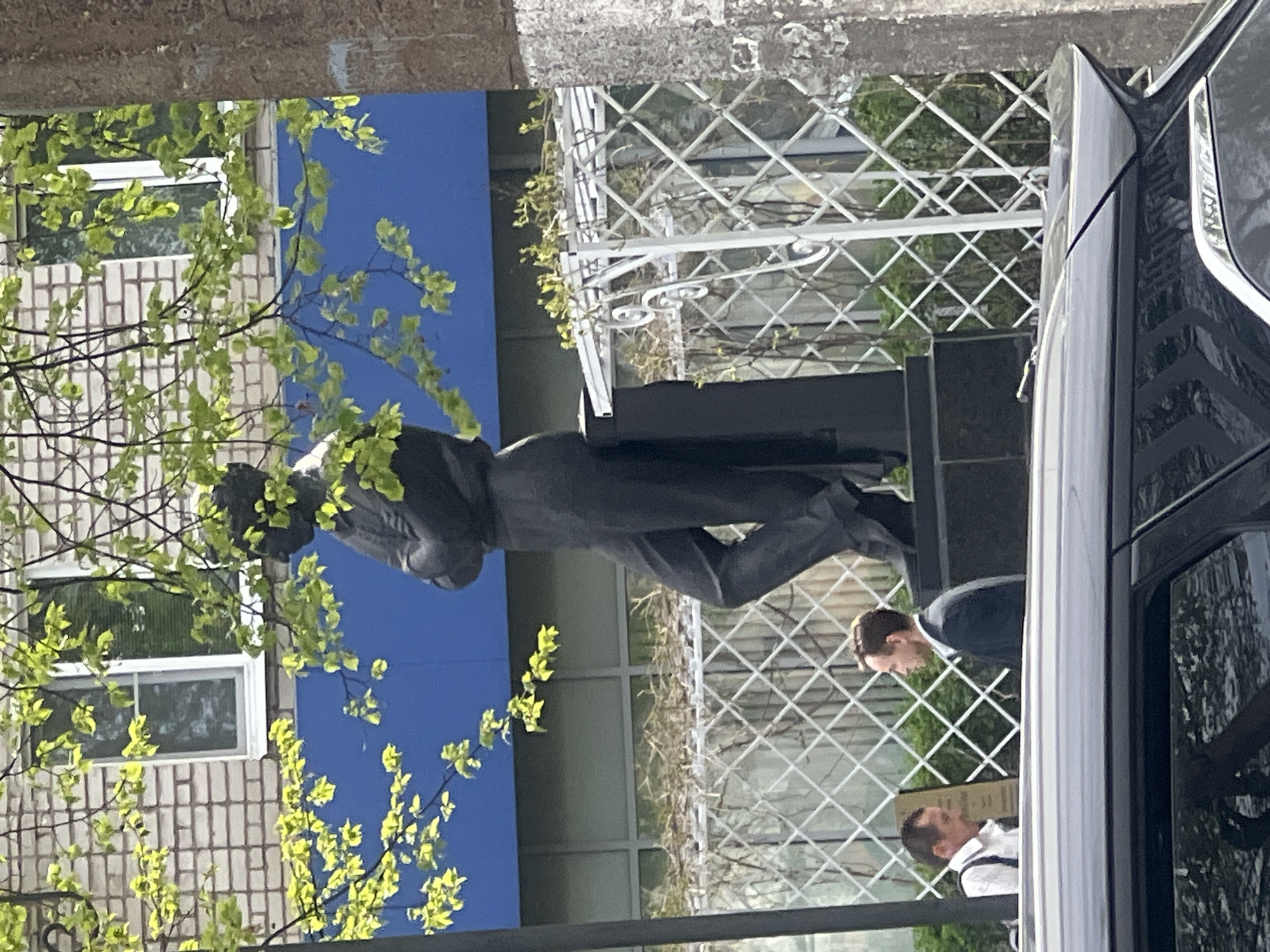 The image depicts a statue in the city of Chaikovsky, Russia, viewed partially obscured by foliage and the side of a dark-colored vehicle. The statue is of a man, possibly leaning slightly, dressed in what appears to be a coat or suit jacket with trousers. It stands on a dark-colored base. Behind the statue is a light blue wall or facade, a section of a light-colored brick building with a window, and a white, geometric-patterned fence. Two men, one in a dark suit and the other in a white shirt with suspenders, appear to be looking at or interacting with the statue. The composition suggests the photo was taken from a street level.