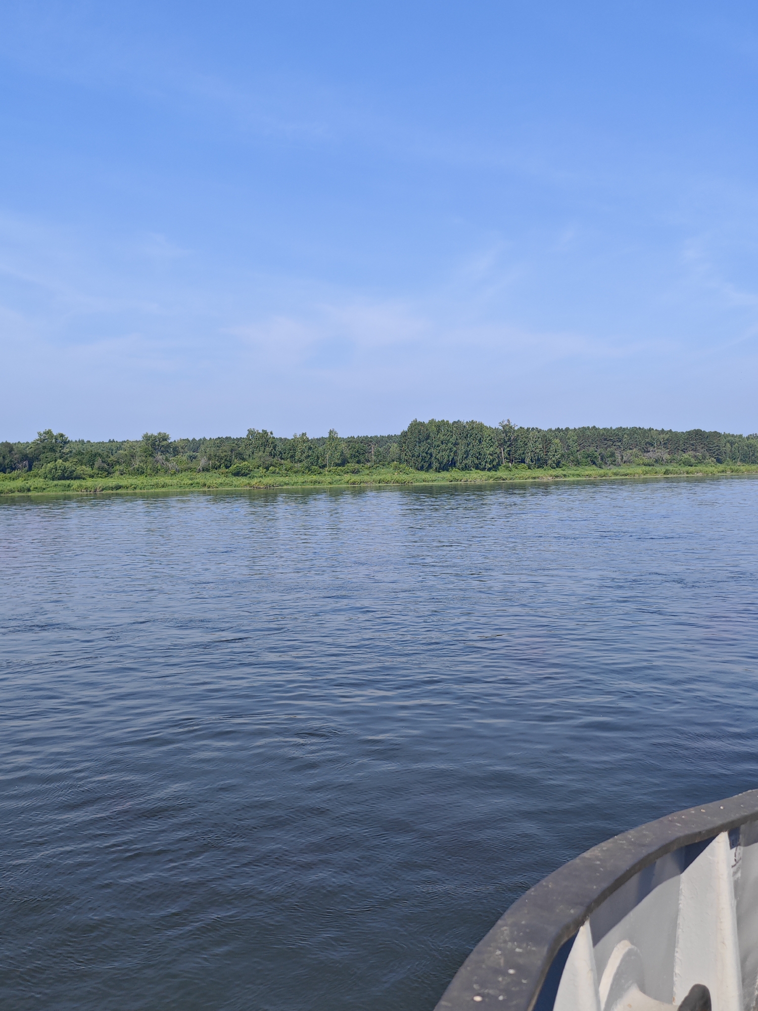 A wide, dark blue river flows towards a densely forested bank under a clear blue sky with wispy white clouds. The water is slightly rippled, suggesting a gentle current. The forest on the opposite bank is a lush green, with various shades indicating different types of trees and foliage. In the foreground, the edge of a boat is visible, with a dark grey railing and a light grey interior. The boat appears to be moving, as indicated by the perspective and the ripples in the water. The scene is peaceful and natural, likely taken from the perspective of someone on a boat on the river near Samkovo, Russia.