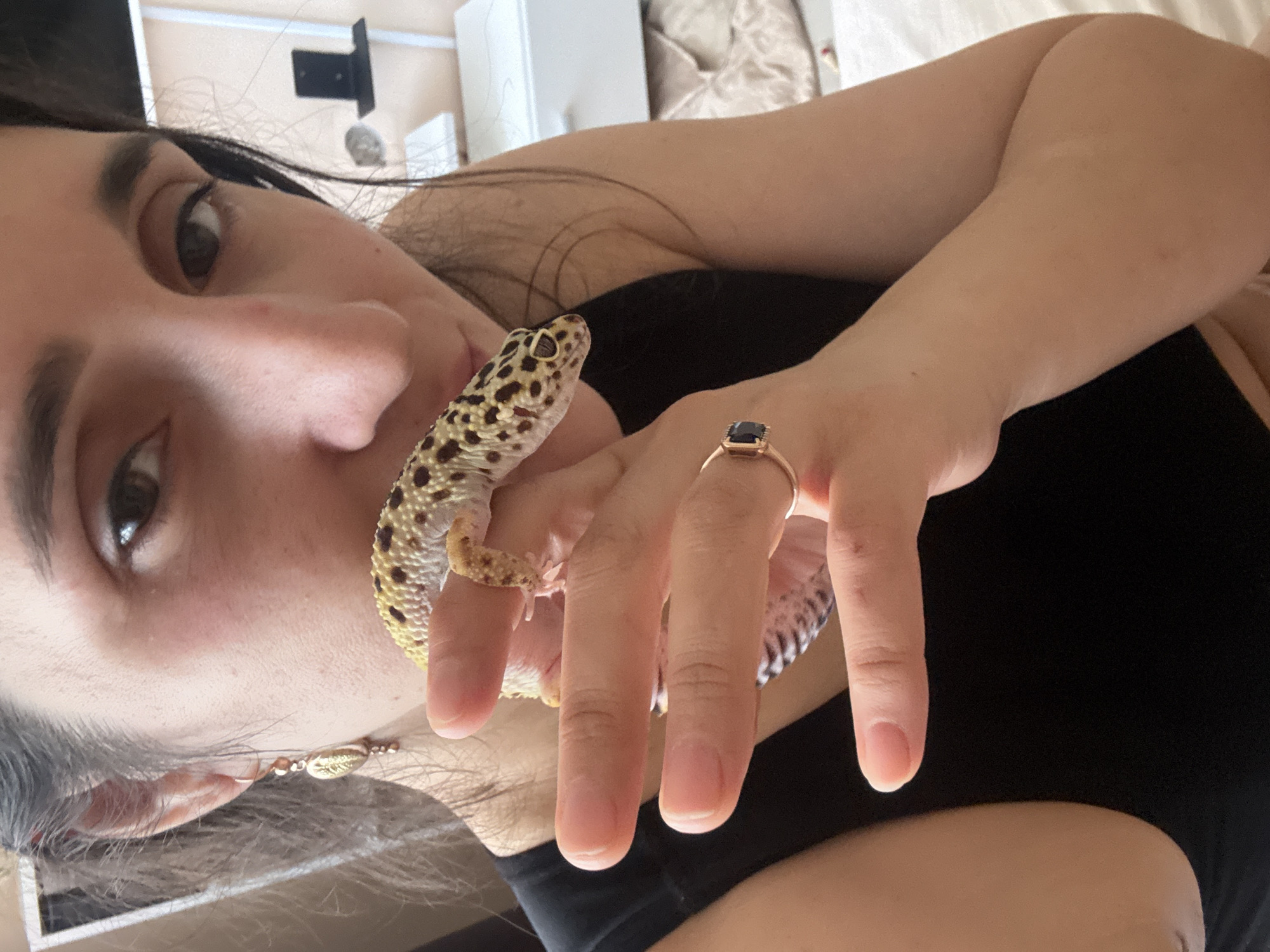 A woman holds a leopard gecko on her hand, close to her face. She wears a dark shirt, earrings and a ring. The background is blurry, showing furniture. There is no indication of the location being Tobolsk, Russia.