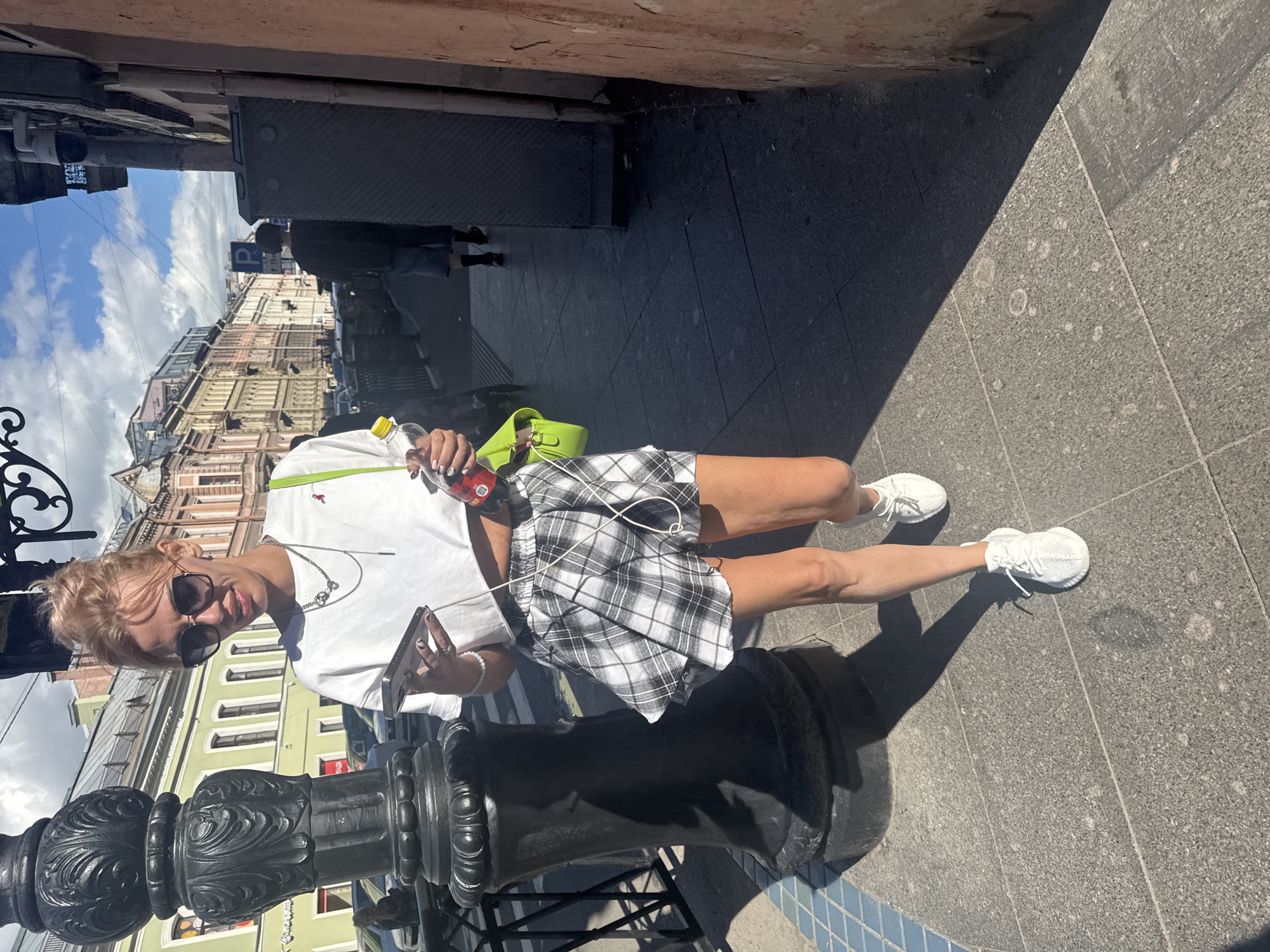 A woman with blonde hair is standing on a sidewalk in Saint Petersburg, Russia. She is wearing sunglasses, a white t-shirt, a black and white plaid skirt, and white sneakers. She is holding a bottle of what appears to be a beverage in her right hand and a smartphone in her left hand. She has a lime green purse slung over her shoulder. A dark metal ornate lamppost is in the foreground. The background shows buildings with traditional European architecture and a cloudy blue sky. The sun is casting a strong shadow, indicating it is likely daytime. The woman appears to be looking at her phone and smiling slightly. The overall scene suggests a casual walk on a sunny day in an urban environment. There is no visible text in the image.