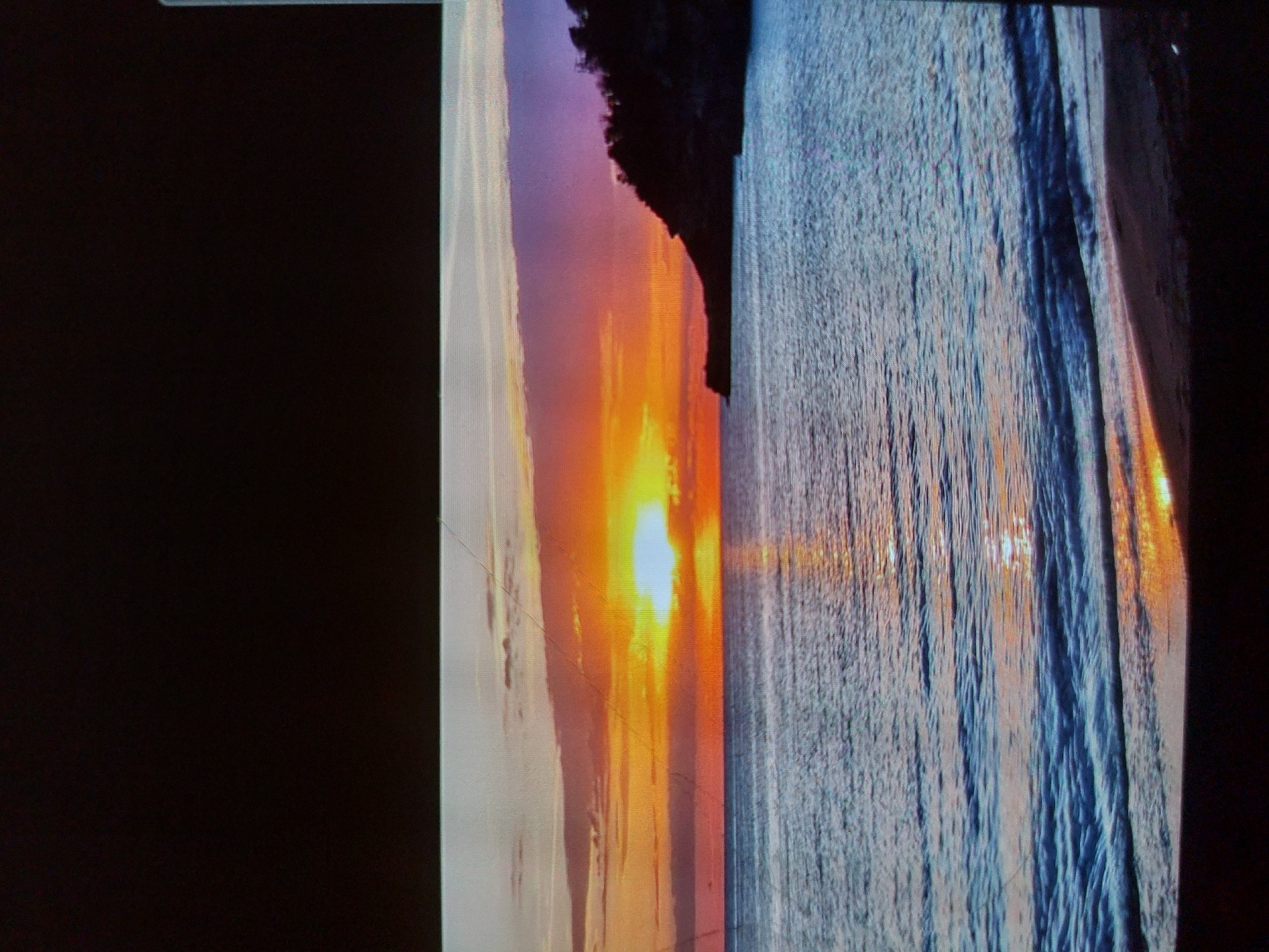 This media file captures a sunset over a calm sea, featuring vibrant colors and a dramatic natural landscape. The image is a photograph of a screen, showing noticeable pixelation and faint horizontal lines across the display.
**Scene and Setting:**
The central focus is a brilliant sunset over a large body of water, likely an ocean or sea. The sky transitions from an intense, almost white-hot yellow around the sun, spreading into rich oranges, reds, and pinks at the horizon, and fading to soft purples and light blues higher up. A dark, silhouetted landmass, possibly a cliff or coastal hill, frames the left side of the scene, standing starkly against the brightly colored sky.
**Subjects:**
The primary subjects are the sun, the expansive sky, the water, and the silhouetted coastline. The sun is a powerful, radiant orb low on the horizon. The water exhibits a textured surface with ripples and small waves, reflecting the dynamic colors of the sky and creating a shimmering path of light from the sun towards the viewer. No people, animals, or man-made objects are visible, emphasizing the untouched natural beauty of the moment.
**Activity or Event:**
The image captures the natural event of a sunset, a daily occurrence known for its visual splendor.
**Notable Details:**
The time of day is clearly sunset, as indicated by the position and color of the sun and sky. The weather appears clear and calm, with no clouds obscuring the sun and gentle movement on the water's surface. The dominant emotions evoked are serenity, awe, and perhaps tranquility. The location context suggests this scene could be along the coast of Tsarevo, Bulgaria, though no specific landmarks are identifiable in the image itself. There is no visible text in the image.