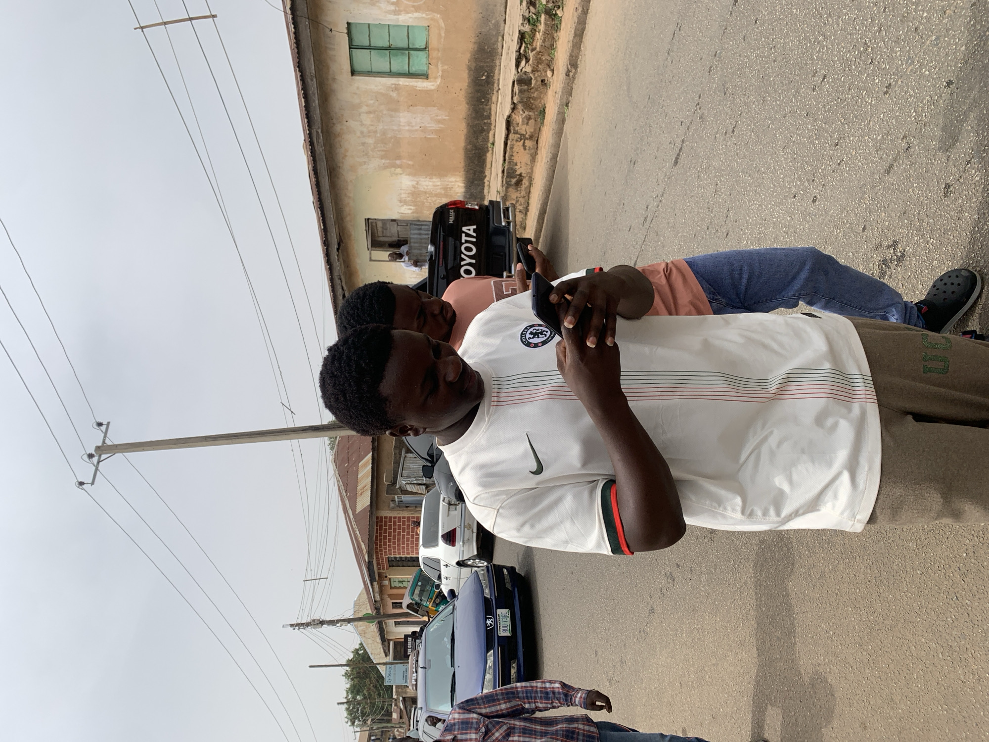 Two young men stand on a street in Jos, Nigeria, both looking at their mobile phones. One wears a white Chelsea football jersey with green and red accents, while the other is in a salmon-colored shirt and blue jeans. Behind them is a Toyota Hilux pickup truck, and several other cars are parked along the side of the road. The buildings are aging and in need of repair. Overhead are power lines. The road surface appears to be asphalt.