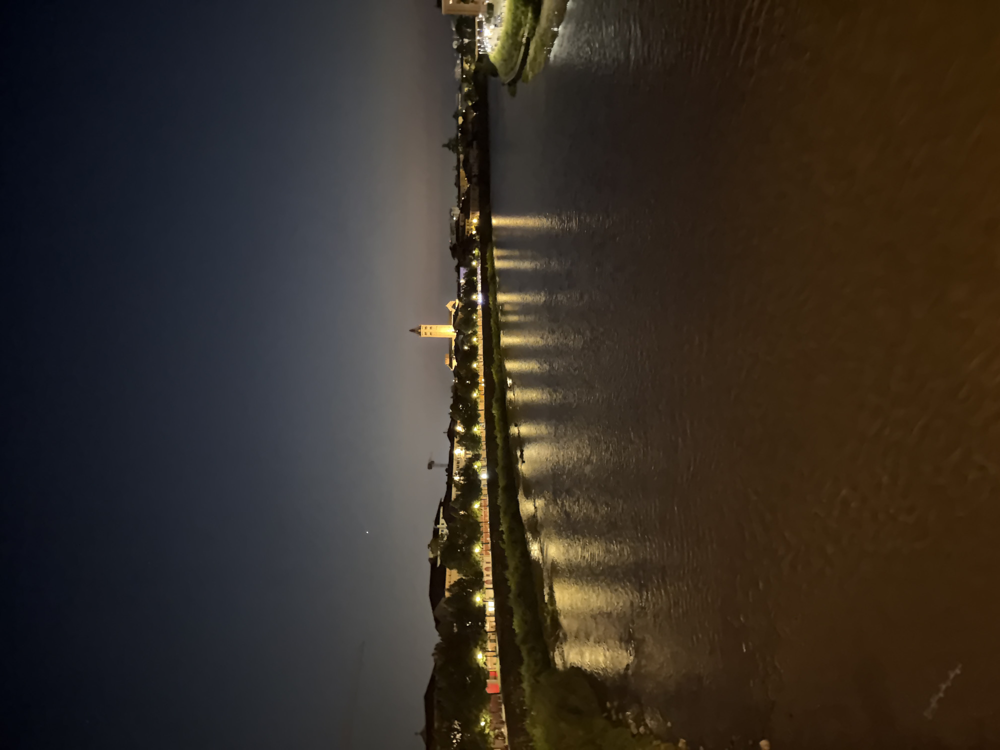 The image depicts a nighttime view of the Adige River in Verona, Italy. The river flows through the center of the frame, reflecting the lights of the city. On the left bank of the river, a row of buildings with illuminated windows and streetlights can be seen, lined with trees. A prominent bell tower, likely part of a church or historic building, stands out against the dark sky in the distance. The right bank of the river appears to be a landscaped area with some lights visible. The sky is dark blue, with a hint of twilight fading. There are no people visible in the image. The overall scene conveys a serene and picturesque urban landscape at night.
