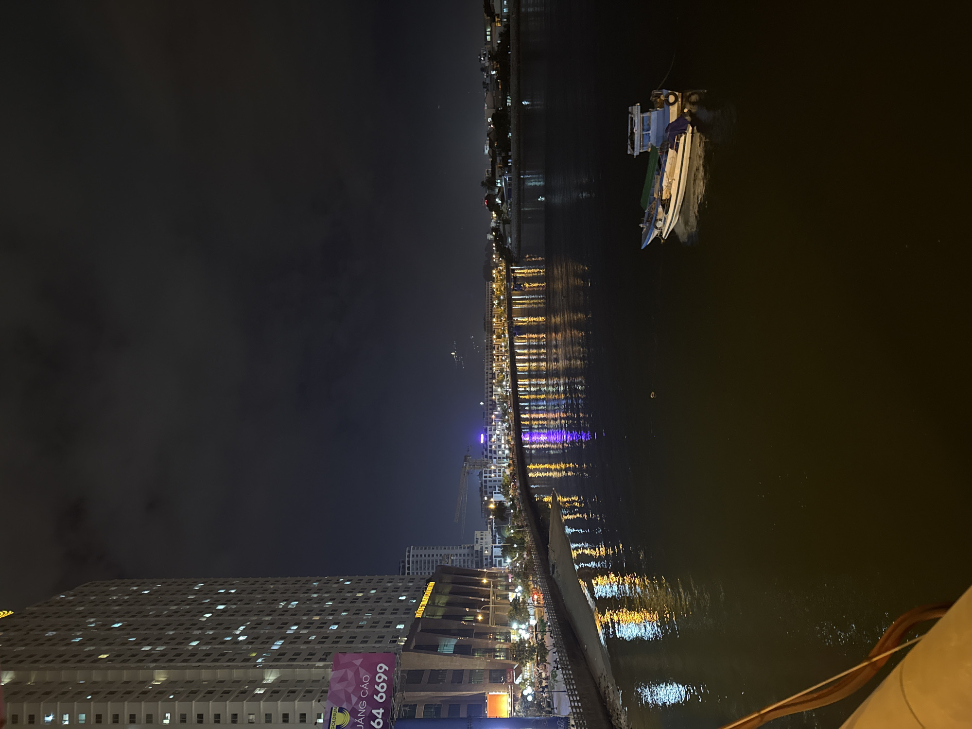 The image depicts a nighttime scene along a waterfront in Nha Trang, Vietnam. On the left, a tall, modern apartment building dominates the foreground, its windows illuminated by warm light, indicating occupancy. Adjacent to the building is a darker, multi-story structure with large windows and architectural details. A billboard with Vietnamese text and numbers is partially visible on the left side.
The waterfront promenade features streetlights casting reflections on the dark water of the river or bay. A sandy area or small beach runs along the edge of the water. Across the water, the city skyline is visible, characterized by numerous buildings of varying heights and illuminated windows, creating a vibrant urban glow. Several boats are present on the water. In the mid-ground, a string of lights from the distant shore reflects on the water's surface, creating a shimmering effect. In the foreground, a small, light-colored boat with a blue cabin and a green tarp is anchored in the dark water. The sky is dark and cloudy, typical of nighttime. The overall impression is of a bustling city at night, with modern architecture and vibrant nightlife.