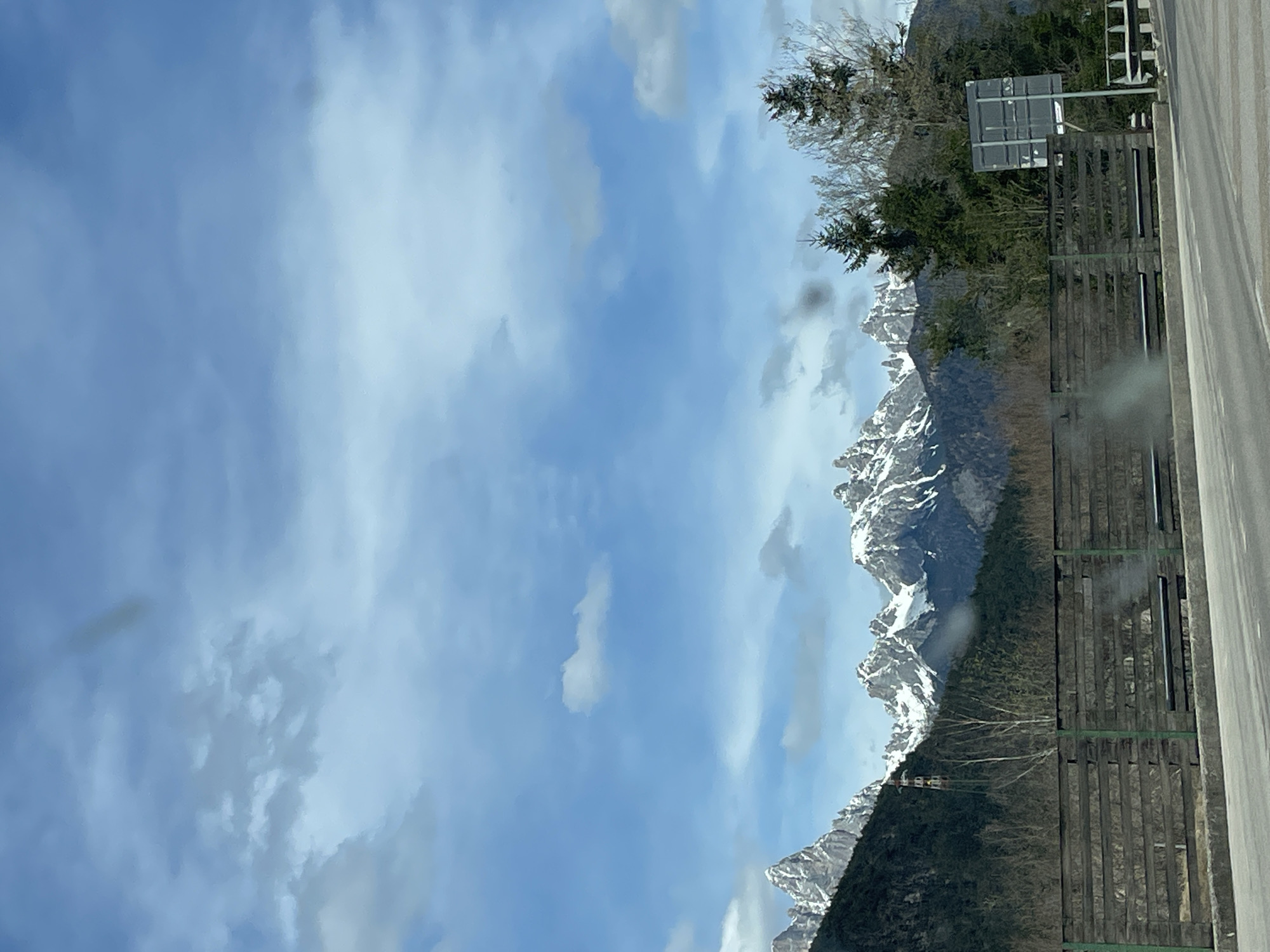 The image depicts a mountainous landscape under a partly cloudy, blue sky. A range of snow-capped mountains is visible, descending into a forested area. A wooden fence runs alongside what appears to be a road or pavement. A sign with a grey surface is attached to a post, adjacent to the fence. The location is Caralte, Italy.