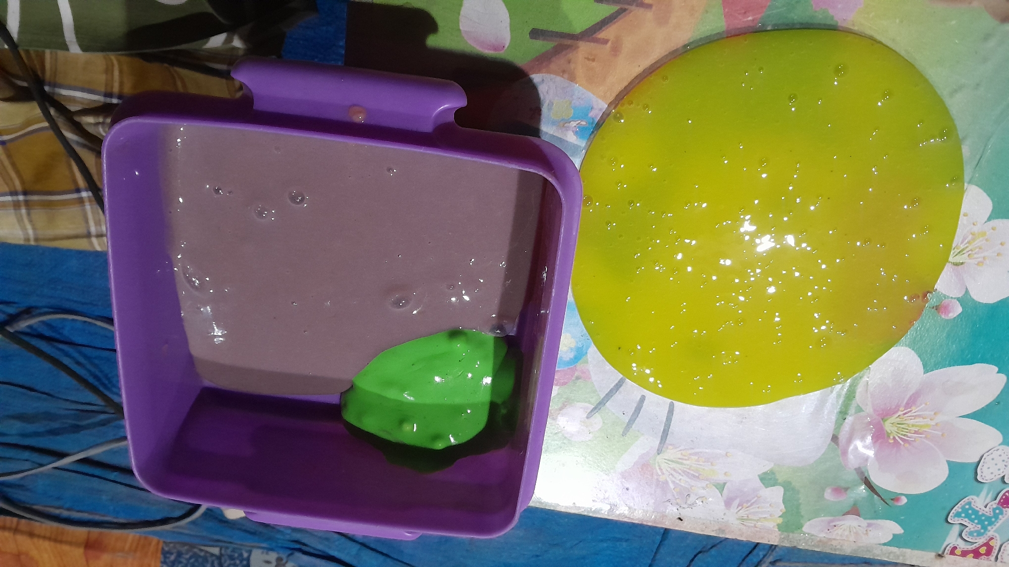 A close-up, overhead shot shows two containers of slime. On the left, a flat, circular mass of bright yellow slime glistens with tiny air bubbles, resting on a colorful surface patterned with delicate pink and white flowers. To the right, a purple rectangular plastic container holds a portion of purplish-brown slime with a blob of bright green slime on top. The container itself has a slightly reflective, glossy finish. The background consists of a blue tarp and a brown and tan plaid fabric, suggesting an indoor setting, possibly in Sumberarum, Indonesia.