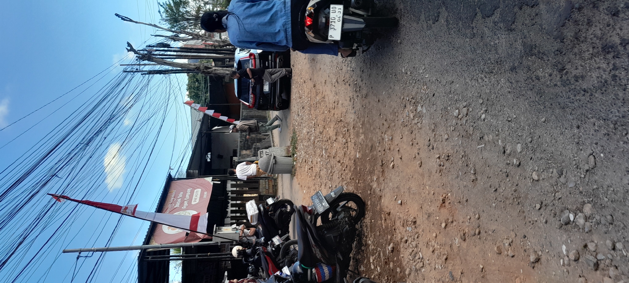 The image depicts a street scene in Rogojampi, Indonesia, on a sunny day. In the foreground, a dark-colored motorcycle with a license plate reading "P 3710 UO" is parked. The rider, wearing a blue denim jacket, is partially visible from behind. To the left, another motorcycle is parked, and behind it, a woman in a white shirt and headscarf is standing.
Further down the street, a black SUV is parked. Several people are in the vicinity of the SUV. One man in a white t-shirt and light-colored pants is walking away from the viewer. Another man in a brown jacket and light-colored pants is holding a white object and appears to be interacting with someone inside the SUV. A large banner with red and white stripes and Indonesian text is displayed on a building on the left. There are numerous overhead electrical wires crisscrossing the sky. The ground is a mix of asphalt and dirt with scattered rocks. The environment suggests a commercial or residential area with a slightly unkempt appearance.