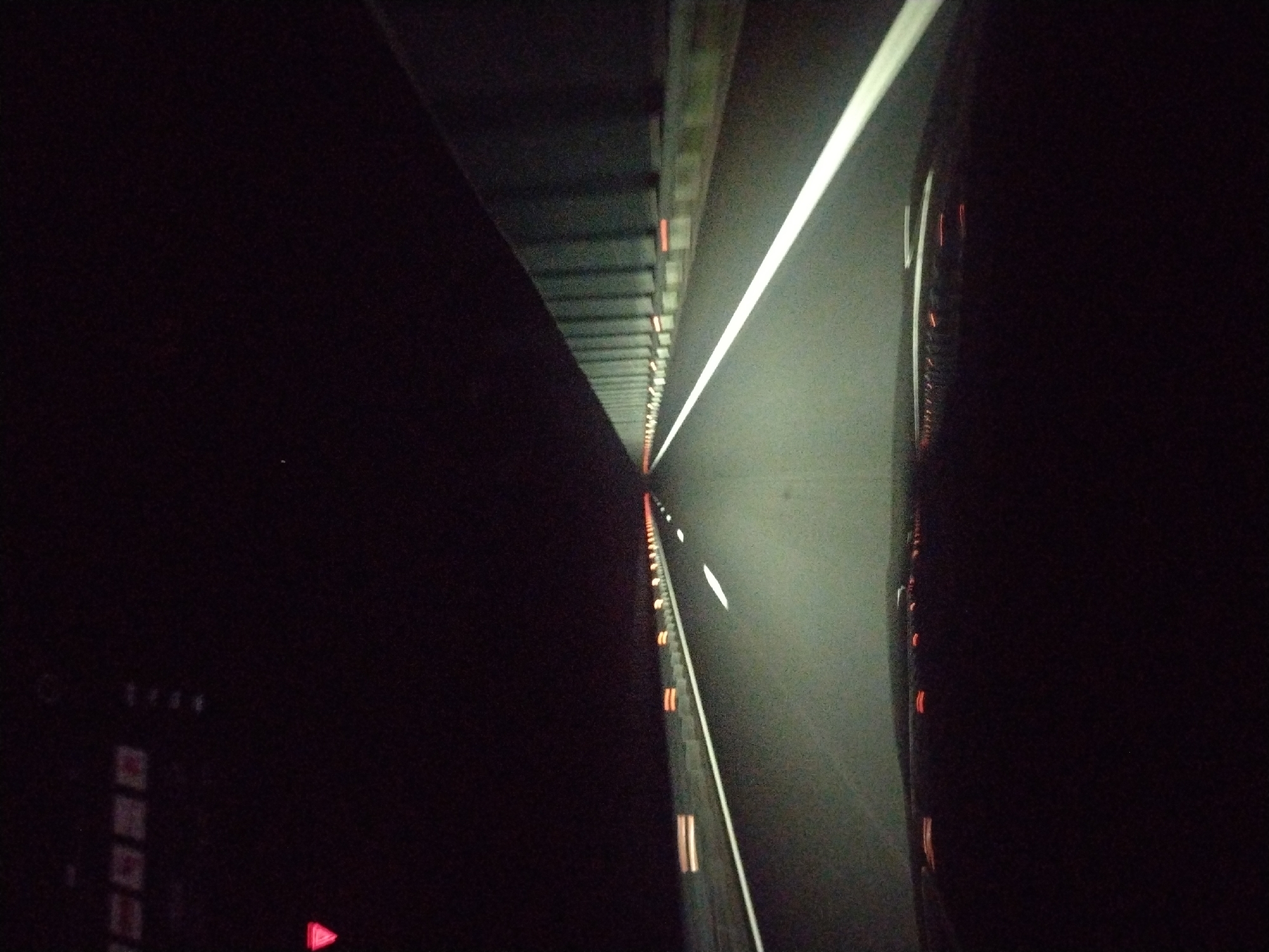 The image depicts a dark highway at night. A long, straight stretch of asphalt is illuminated by the headlights of a vehicle, possibly a car, from which the image was taken. A bright white line marks the edge of the lane. On the left side, a structure runs parallel to the road, with periodic orange lights. The dashboard of the vehicle is visible in the lower left corner, showing faint indications of the car's instruments. Due to the darkness and lack of recognizable features, the specific location within Chokoba, Bulgaria cannot be determined.