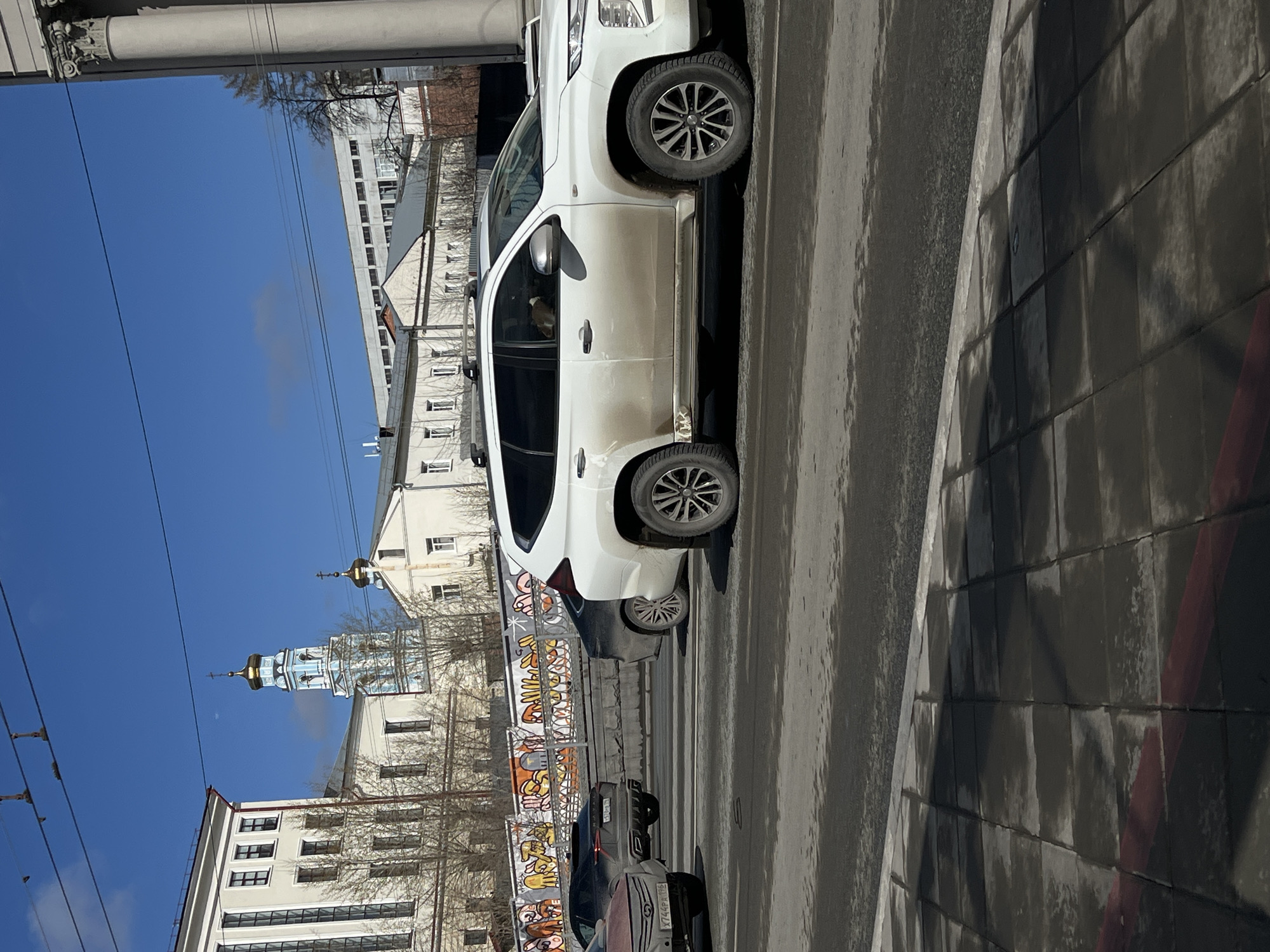 The image shows a street in Yekaterinburg, Russia, featuring a white SUV parked near a crosswalk. Several other cars are visible in the background. A building with a traditional Russian Orthodox church steeple can be seen in the background. Graffiti decorates a barrier along the street. Overhead power lines are visible against a blue sky. The street and sidewalk appear wet.