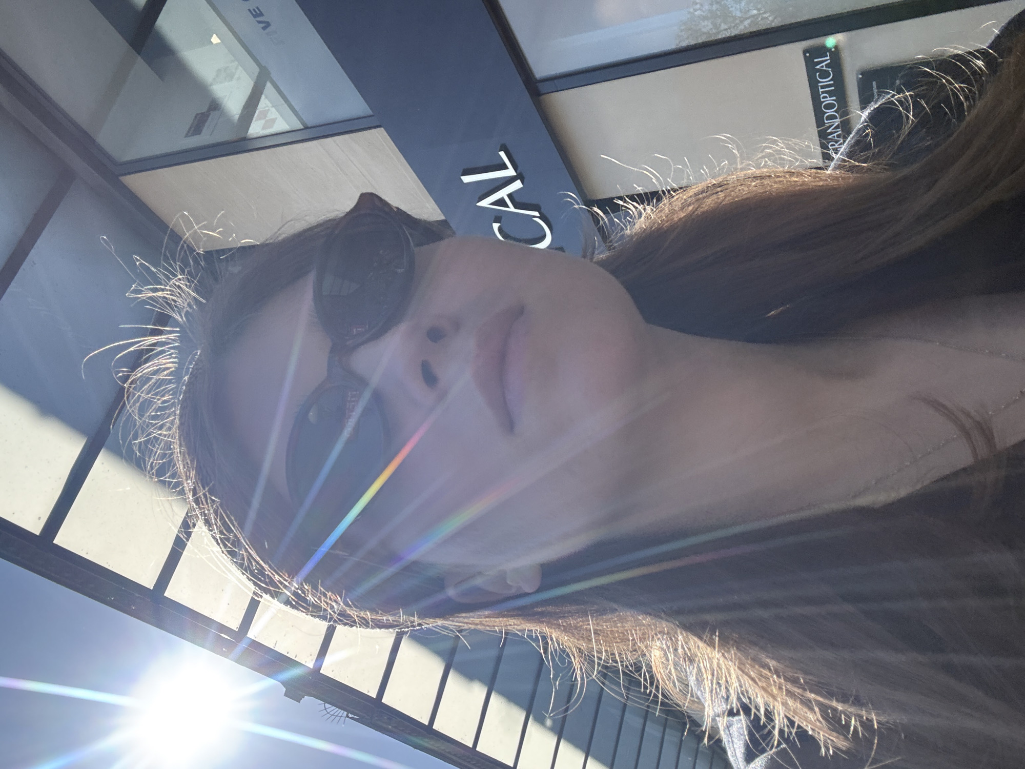 A woman is pictured in an upward angle selfie, wearing sunglasses. The background shows an outdoor environment with a commercial storefront. The sign reads "OPTICAL" and "GRAND OPTICAL." The environment is brightly lit with strong sunlight, creating lens flares. It cannot be determined that this image was taken in Paris, France.