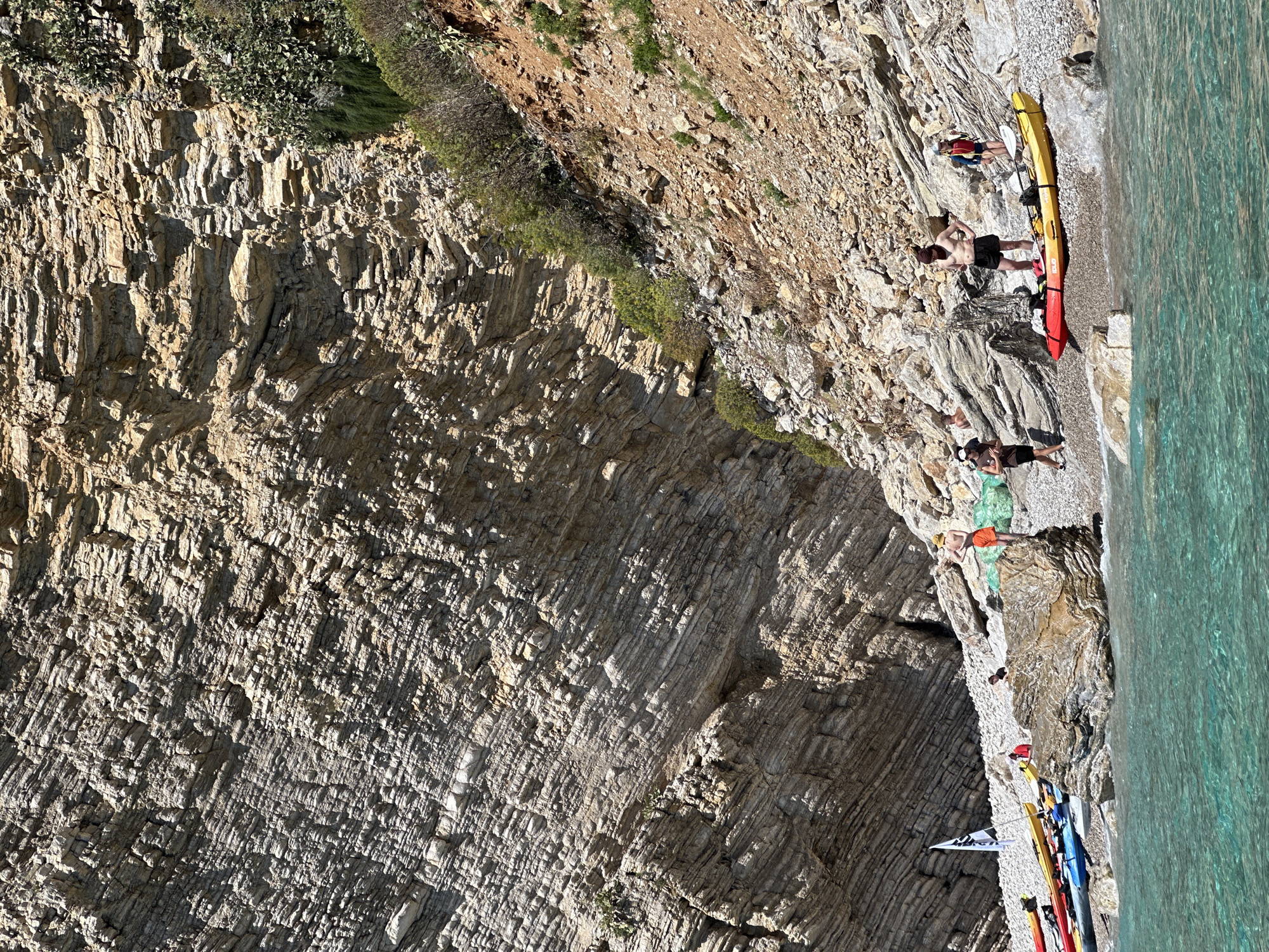 The image captures a scene on a rocky beach, likely in Budva, Montenegro, given the presence of distinctive layered rock formations. The beach is not sandy but composed of small pebbles. Several people are scattered along the narrow strip of beach between the rock face and the sea.
At the top right, a man is standing shirtless near a long, red and yellow kayak. Another person is nearby wearing a red and blue top and shorts.
Further down the beach, another person wearing a green top is sitting with their legs extended towards the sea. An individual wearing an orange top is next to them.
At the bottom right, a collection of kayaks of different colors, including yellow, blue, and red, are lined up on the pebbles. A white flag is partially visible among the kayaks.
The sea is a clear turquoise, indicating calm conditions. The rock face comprises light-colored, horizontally layered rock strata. Vegetation is sparsely distributed along the top of the rock face and in crevices. The overall lighting suggests a sunny day.