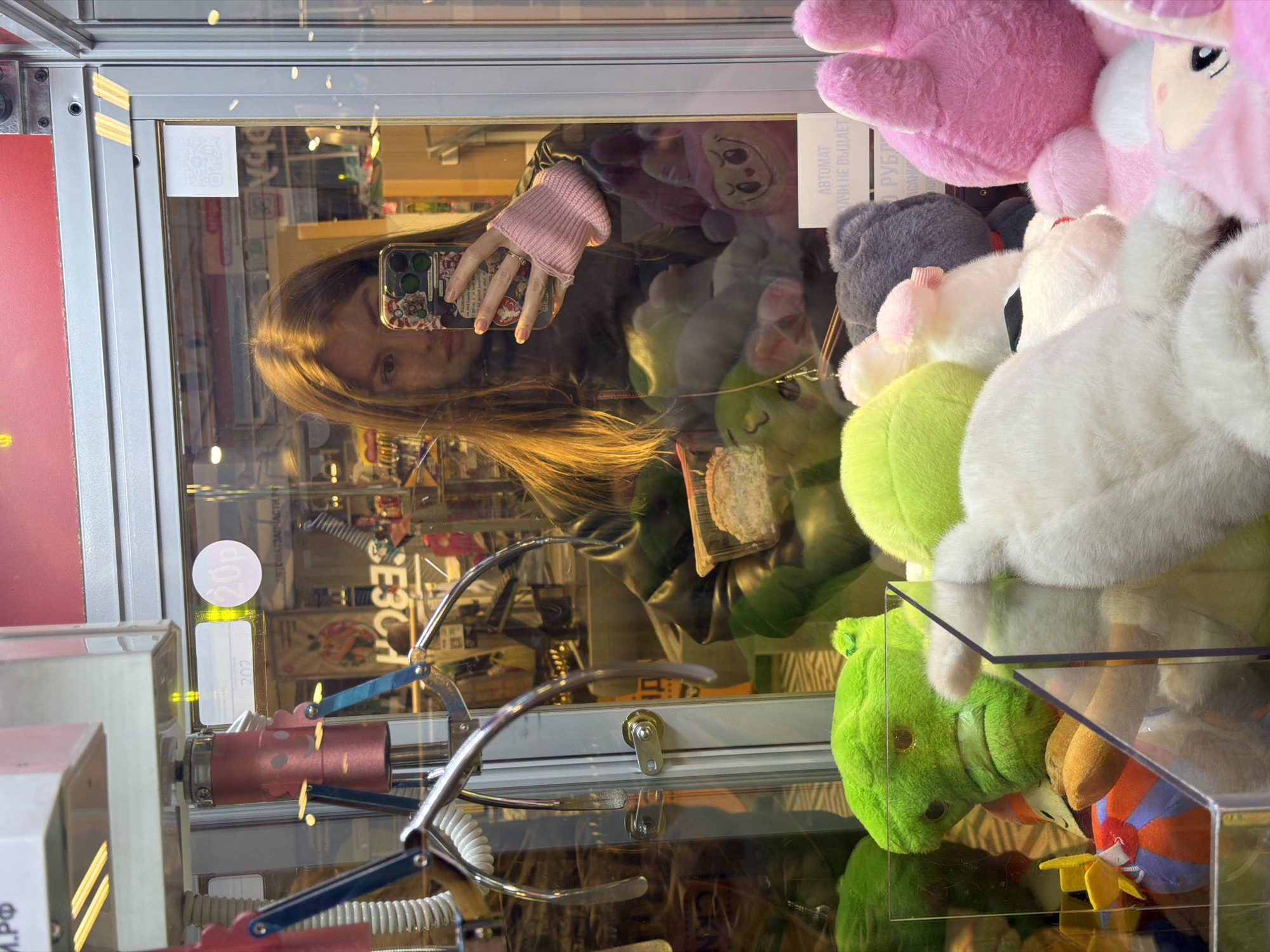 A young woman is reflected in the glass panel of a claw machine, holding a smartphone as if taking a selfie or recording. Her face and part of her upper body are visible, with long, light brown hair falling over her shoulder. She wears a dark jacket and a pink knitted wrist warmer or glove on her left hand, which holds the phone. The phone case has a patterned design.
The scene takes place indoors, likely within an arcade, shop, or shopping center in Khimki, Russia. The claw machine is filled with various plush toys in different colors, including pink, white, green, and grey. Some toys are distinctly shaped, like a green, possibly dinosaur or dragon-like figure, and a pink character with a face. The mechanical claw of the arcade game is visible in the lower-left section of the image.
Reflected in the glass behind the woman and the toys are elements of the surrounding environment, suggesting shelves or displays of other products. Notable details include a small white sticker with a QR code on the upper left, and another sticker inside the machine partially showing Cyrillic text. Visible words include "АВТОМАТ" (AUTOMAT) and fragments like "РУБЛЕЙ" (RUBLES) and "НЕ ВЫДАЁТ" (DOES NOT ISSUE), indicating that the machine is an automated prize dispenser that accepts rubles but does not dispense cash. Another small round sticker reads "20p" or "20k". The overall lighting suggests a well-lit indoor environment.