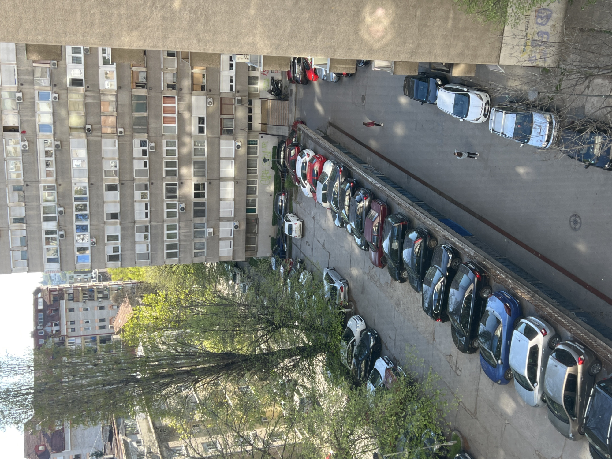 An overhead shot reveals a cityscape in Belgrade, Serbia. In the foreground, a large, pale apartment building with numerous rectangular windows and air conditioning units jutting out. A line of parked cars runs along a concrete walkway adjacent to the building, a diverse array of colors and models. Trees with budding green leaves fill the lower left of the frame. A few people walk near parked cars. In the background, another building with brown roofs and light-colored walls, partially obscured by trees. The overall lighting suggests a sunny day.