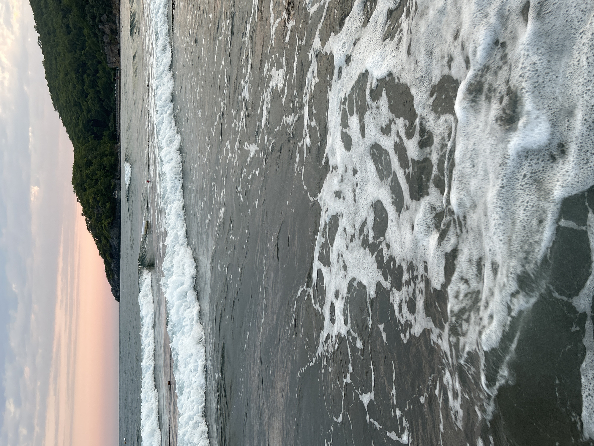 The image shows a view of Iztuzu Beach in Ortaca, Turkey, during what appears to be sunset or sunrise. The foreground is dominated by the dark, foamy water lapping at the shore, with white foam forming intricate patterns on the surface. Two distinct waves, one in front of the other, are breaking. In the mid-ground, the sea stretches towards a lush, green, tree-covered hill that forms the backdrop. The sky above is a soft gradient of pale pink and grey clouds. There are no people or specific objects clearly visible in the water or on the beach that indicate any activity. The overall impression is of a serene natural landscape.