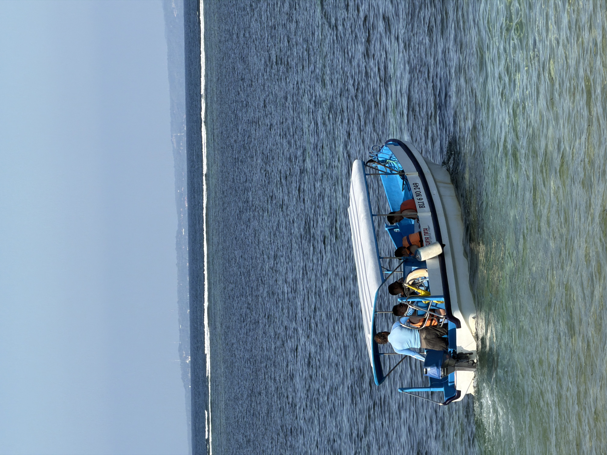 A small, white motorboat with a blue trim and canopy is cruising on clear, turquoise water, generating a foamy wake. The boat is occupied by at least five individuals, including a person in a light blue long-sleeved shirt steering the vessel, and several passengers wearing life vests. One passenger is visible in an orange life vest, and another is partially obscured. Text on the side of the boat reads "BLI 6 NO 249" and "SURYA TIRTA". In the background, a distant landmass with greenery and structures is visible, separated from the foreground by a dark blue expanse of deeper ocean and a line of white-capped waves. The sky is a pale, clear blue. The image was captured in the waters off Denpasar, Indonesia, likely near a popular tourist area due to the presence of recreational boats.