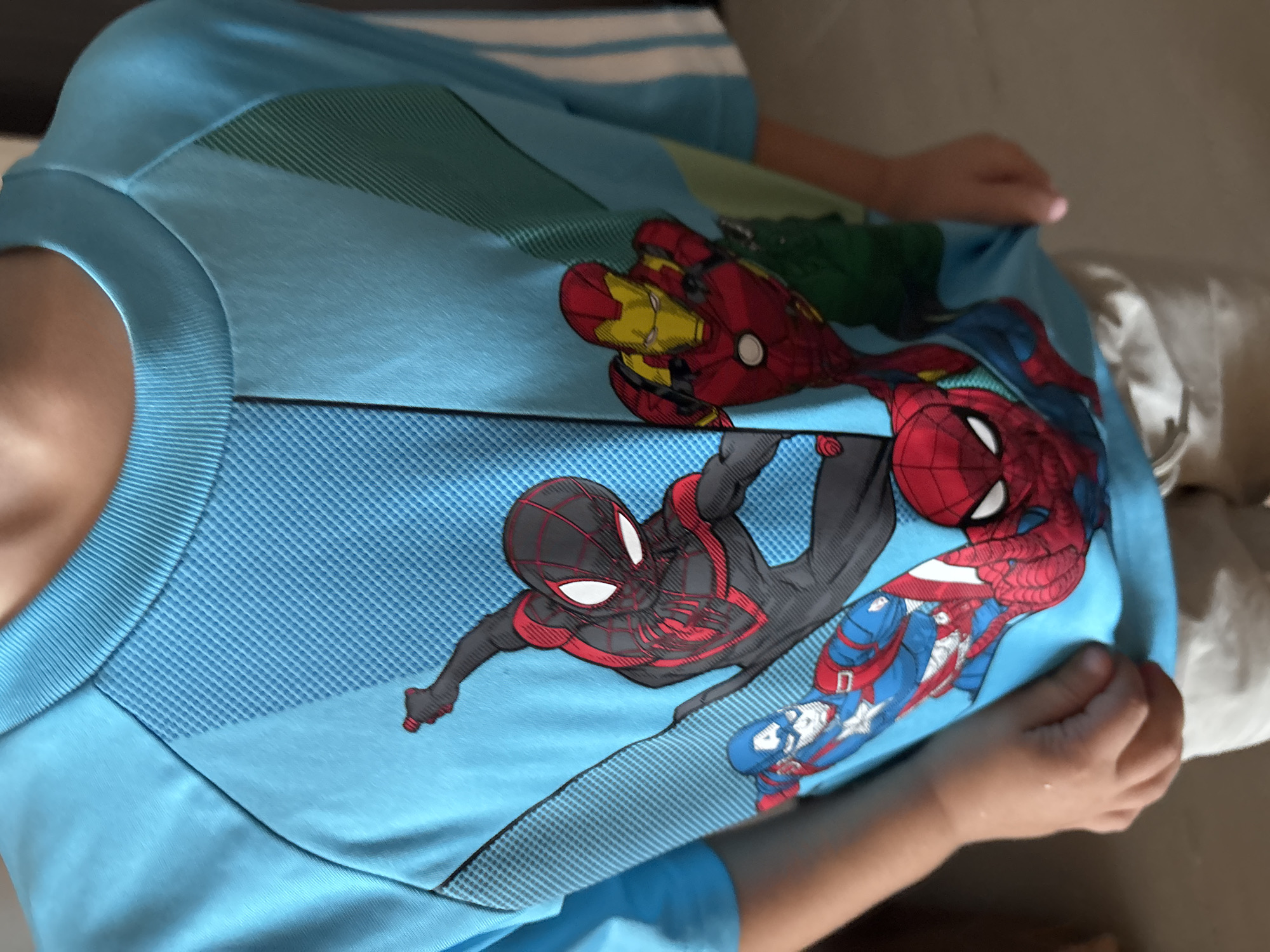 A young child is wearing a bright blue t-shirt featuring a graphic print of Marvel superheroes. The print includes Spider-Man, Captain America, and Iron Man, with Spider-Man prominently displayed in the center. The t-shirt has a crew neck and short sleeves with white stripes on the right sleeve. The child is standing, and the image focuses on the t-shirt and the child's upper body. The background is out of focus but appears to be an indoor setting, possibly a home, with neutral-colored flooring visible at the bottom of the frame. There is no discernible information about the specific location in Dubai, United Arab Emirates, as the image contains no identifying landmarks or contextual clues related to the city.