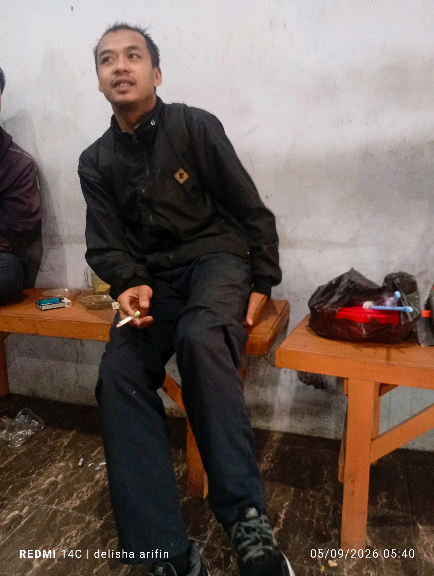 A man with short black hair and a light complexion is sitting on an orange wooden bench. He is wearing a black zip-up jacket with a circular patch on the left breast and dark pants. His right hand is holding a lit cigarette. In front of him on the bench are a pack of cigarettes and a small glass dish containing liquid. Another person, partially visible, is sitting to his left, wearing a dark jacket. To the man's right on the bench is a black plastic bag containing red and blue items. The floor is a dark, patterned material. The background is a textured off-white wall. The image is watermarked with "REDMI 14C | delisha arifin" and "05/09/2026 05:40". The scene appears to be indoors, possibly in a low-lit establishment in Gambir Kuning, Indonesia.