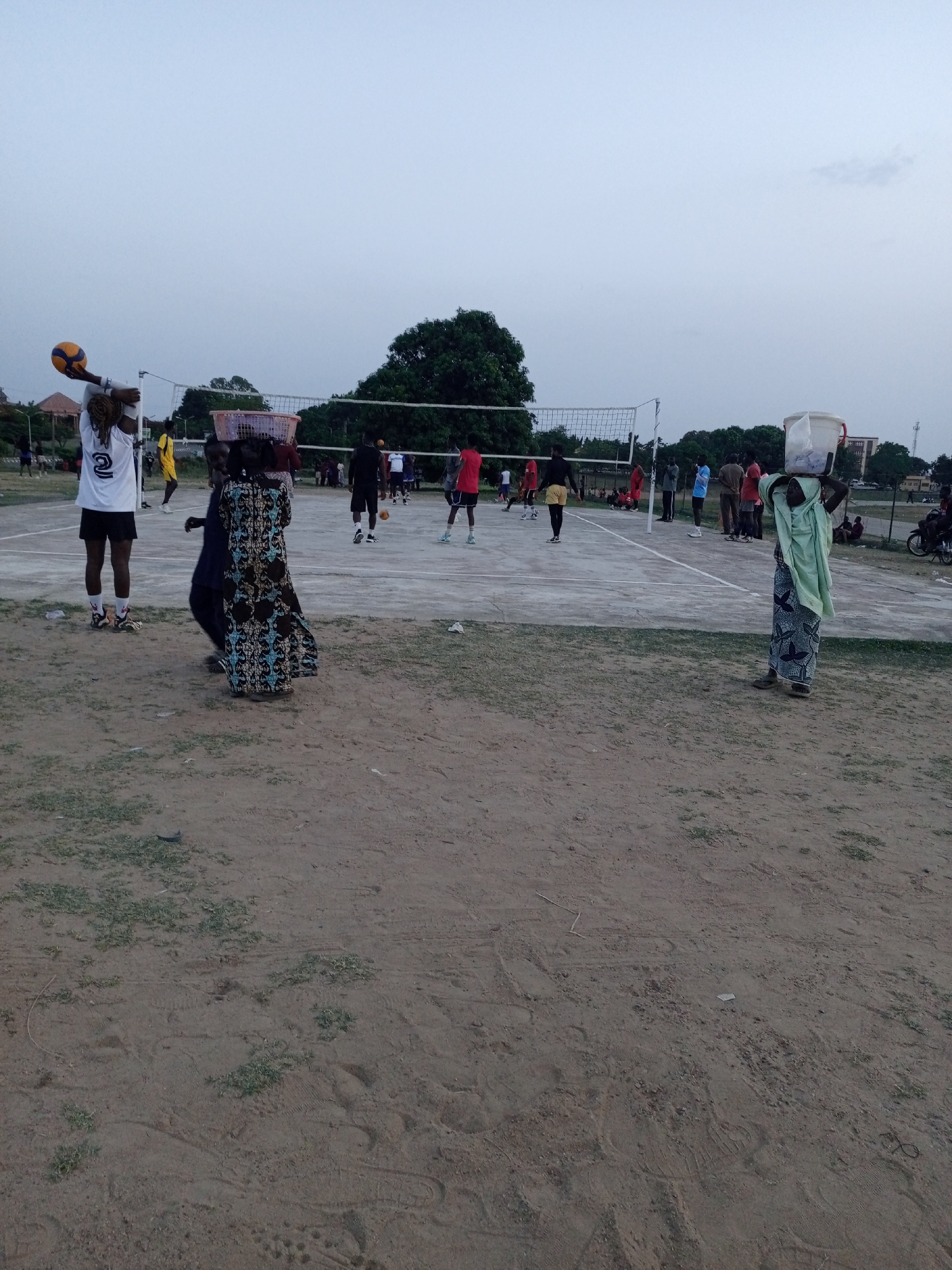 The image captures an outdoor scene in Keffi, Nigeria, featuring a volleyball game and several individuals. The central focus is a dirt or sandy volleyball court with a white net and boundary lines, where multiple people are engaged in or watching a game.
In the immediate foreground, to the left, a person with dreadlocks, wearing a white jersey with the number "2" on the back and black shorts, holds a yellow and blue volleyball overhead, poised for a serve or spike. Next to them, slightly further back, a woman in a long, patterned dress carries a large, light-colored basket on her head. On the right side of the foreground, another woman, draped in a light green headscarf and a patterned skirt, balances a white bucket on her head. Both women appear to be walking or standing near the court sidelines.
On the volleyball court itself, various players in athletic wear, including shorts and jerseys of different colors (red, yellow, blue, black), are scattered across the court, actively playing or observing. More spectators or participants are visible around the court's perimeter.
The ground surrounding the court is primarily sandy earth with patches of sparse green grass. In the background, a large tree with dense green foliage stands prominently. Beyond the tree and the court, a few distant buildings and what appears to be a telecommunications mast or power pole can be seen under a pale, overcast sky. The diffused light suggests it is either late afternoon or early evening. The overall atmosphere appears recreational and social.