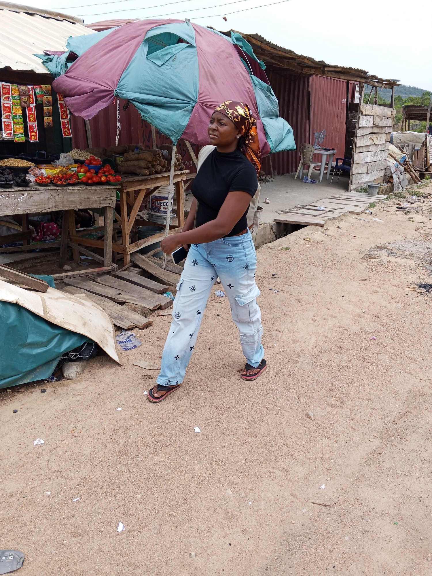 The image depicts a woman walking along a sandy path in what appears to be a market area in Otokiti, Nigeria. The woman is wearing a black top, light blue jeans with patterns, and sandals, and a headscarf. She is holding a phone in her left hand. To her left is a stall selling various food items under a large, patched umbrella. The stall is constructed of wood and displays items like tomatoes, yams, and packaged goods. A blue tarp is partially covering items near the stall. Behind the stall, there's a corrugated metal structure with wooden additions. The ground is sandy and littered with small pieces of trash. A small wooden bridge or platform covers a drainage ditch in the background. In the distance, there is vegetation, suggesting a less urban area.
