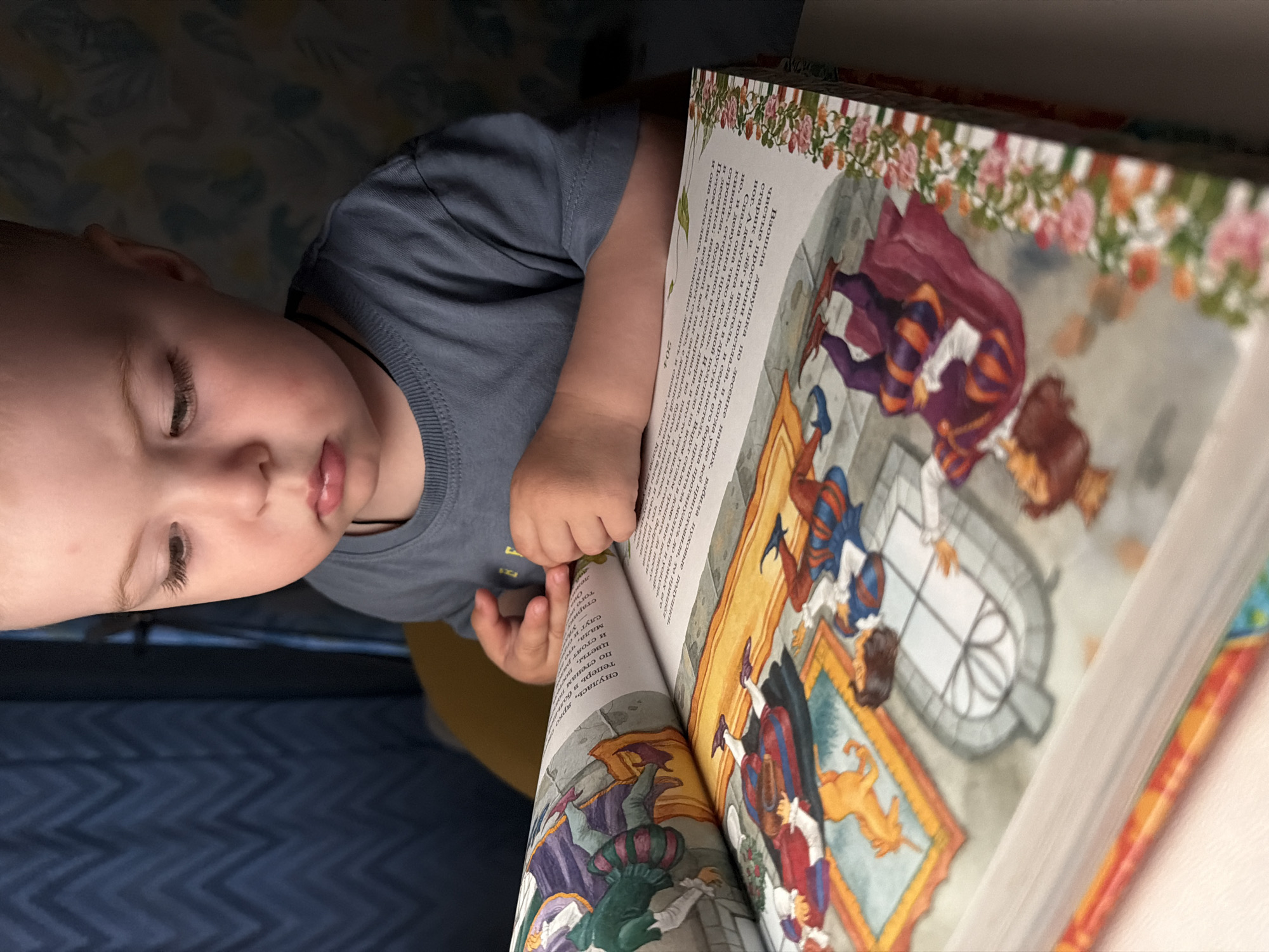 A young child is looking down at an open book placed on a table. The child is wearing a grey t-shirt and has light-colored hair. The book has colorful illustrations and text. The background shows a blue curtain and a wall with a patterned wallpaper. The lighting suggests an indoor setting, possibly a home. The specific location in Votkinsk, Russia, cannot be determined from the image alone.