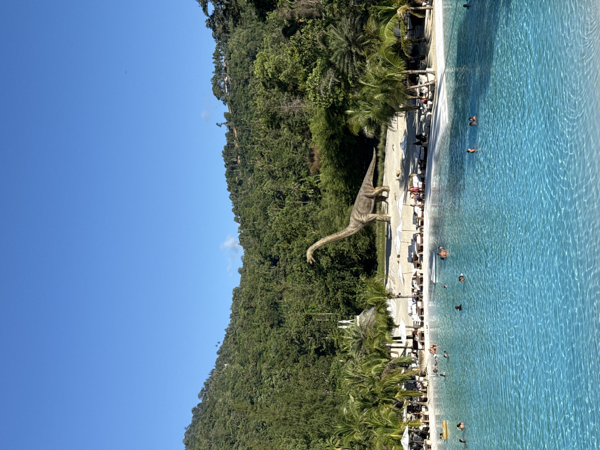 The image depicts a large, vibrant turquoise swimming pool with several people swimming and relaxing. The pool is situated on a sandy beach, lined with lounge chairs and umbrellas, and surrounded by lush green palm trees. In the background, a massive dinosaur statue, resembling a Brachiosaurus, stands prominently on the sand. Beyond the beach and pool area, a dense, tropical forest covers a steep hillside, reaching towards a clear blue sky. The location is the "Namuang Safari Park" in Ko Samui District, Thailand, a tourist attraction known for its natural beauty and the dinosaur exhibit.