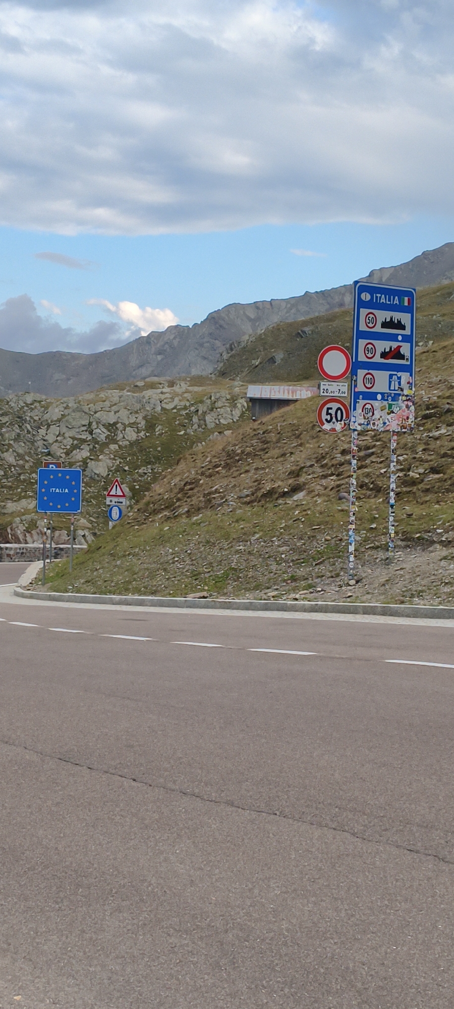 This is an outdoor, eye-level, full shot of a mountain road with multiple signs indicating a border crossing into Italy. In the foreground, a paved road curves to the left with white lane markings. The road surface is a reddish-brown asphalt with a visible crack. To the right of the road, a grassy slope rises towards a rocky mountainside.
On the slope, several signs are visible. A blue sign with the European Union flag and the word "ITALIA" is present on the left. To its right, a triangular warning sign with a red border and an exclamation mark is displayed, followed by a circular sign with a blue background and a white truck symbol. Further to the right, a tall blue sign with "ITALIA" and the Italian flag at the top shows speed limits of 50, 90, 110, and 130 km/h, interspersed with silhouettes of buildings and a train. Below these, a circular sign with a red border and a white circle indicates a restriction, and another circular speed limit sign shows "50" in red. The poles of these signs are covered in numerous stickers. A small, weathered wooden structure with a slanted roof is situated on the hillside behind the signs.
In the background, rugged, rocky mountains dominate the landscape under a partially cloudy sky with blue patches. The overall environment appears to be mountainous and remote, likely a high-altitude pass. There are no people visible in the image. The scene suggests a border crossing point in a mountainous region of Italy, possibly in the Alps.
