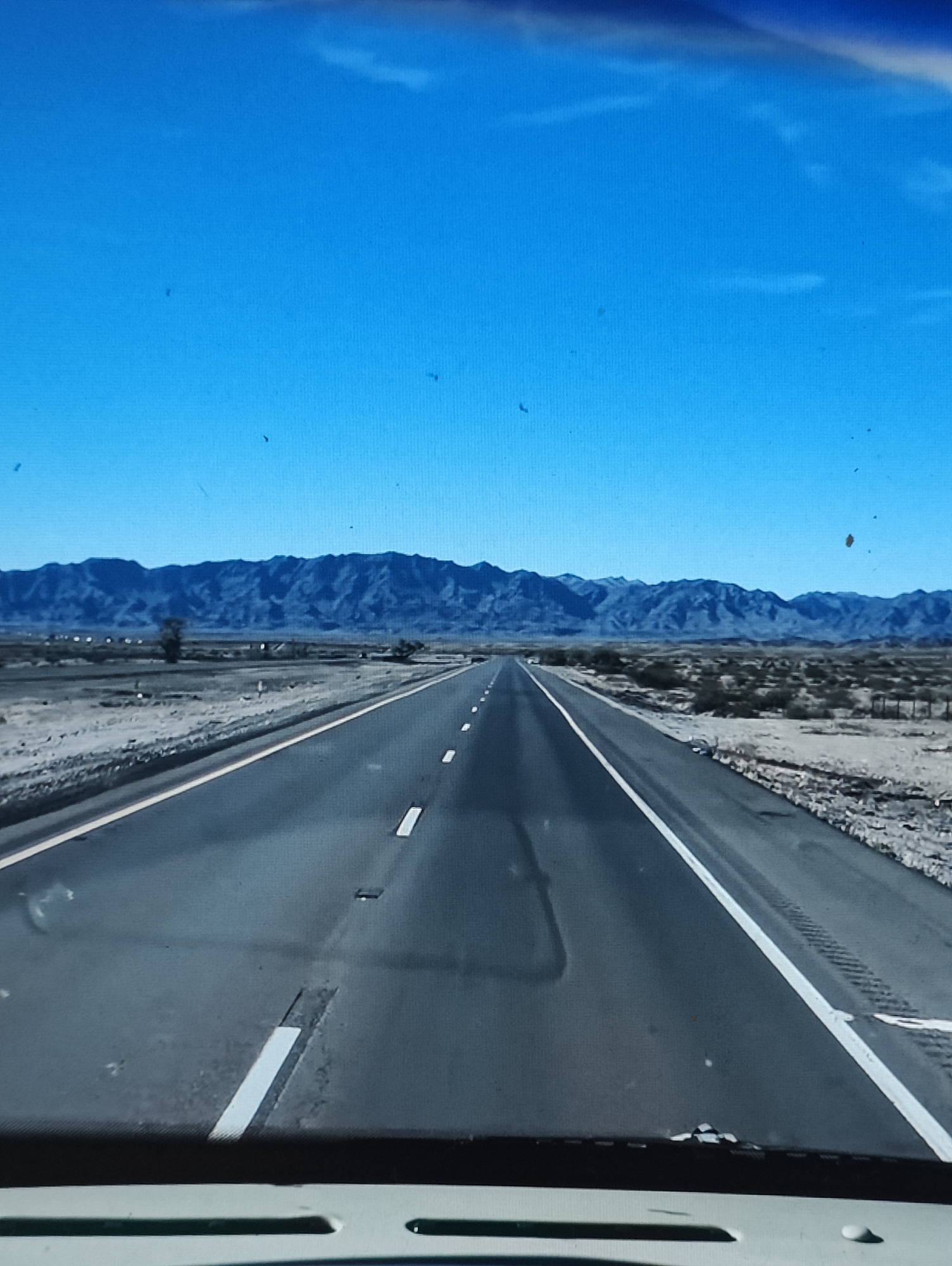 The image shows a view from inside a vehicle looking out at a desert highway stretching towards a mountain range under a clear blue sky. The road has a white dashed line and a solid white line marking the lanes. The desert landscape on either side of the road is arid and sparsely vegetated, with some scrub brush and a few distant trees. The mountains in the background are rugged and appear to be of moderate height. There are no people or animals visible in the image. The scene appears to be in a remote, undeveloped area. The image does not contain any information that would suggest it is located in the city of Yarmolyntsi, Ukraine.