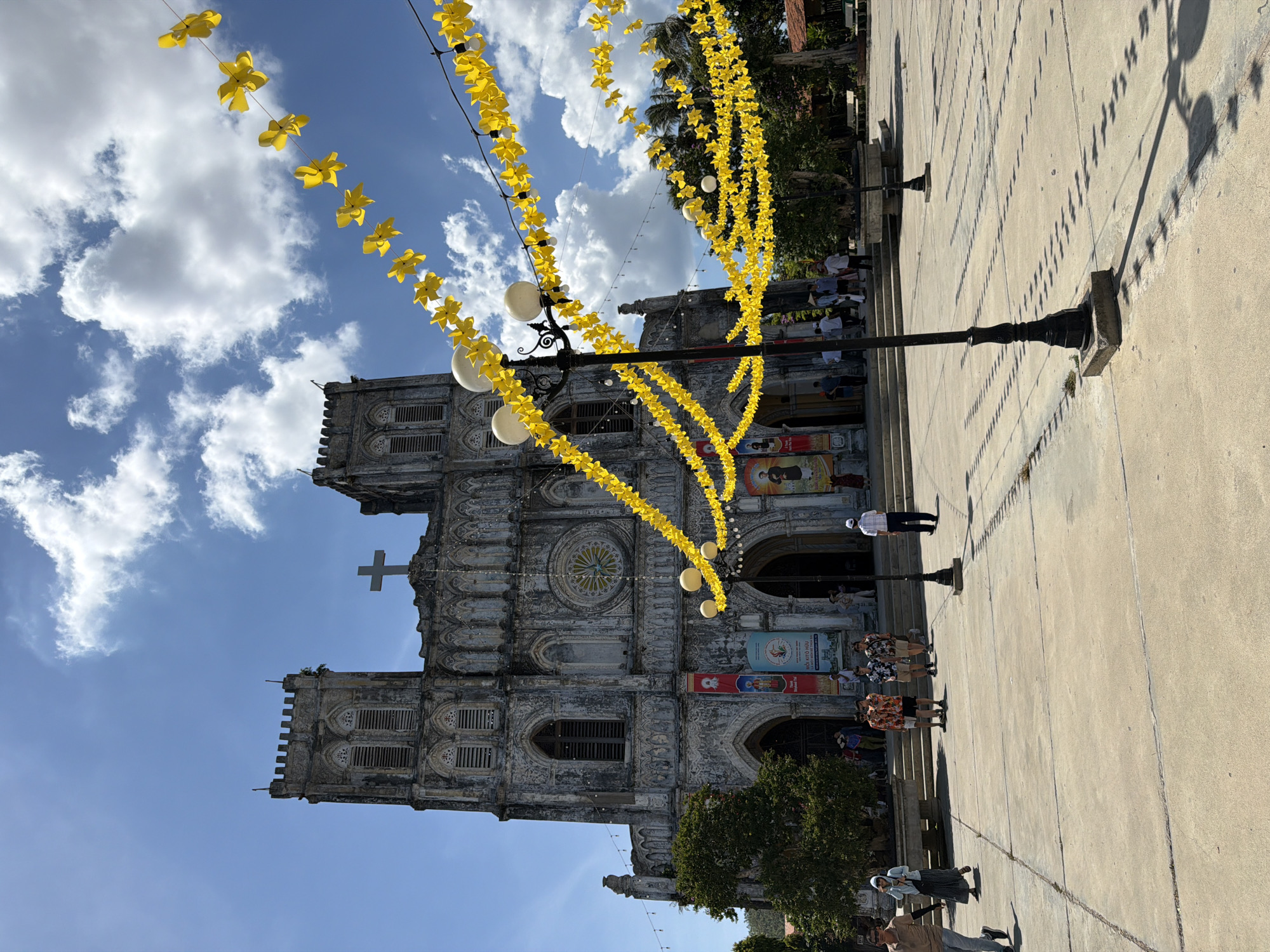 The image captures the exterior of a large, stone church with a weathered facade, characterized by a central rose window and two prominent bell towers. A large cross is visible atop the central section of the building. The church is adorned with numerous strings of yellow, star-shaped decorations that hang across the foreground, suspended by wires and poles. In front of the church, a paved courtyard is visible, with several people walking and standing. Among them are individuals wearing casual clothing, including t-shirts, shorts, and skirts. Some individuals appear to be tourists. Two lampposts with spherical lights are positioned in the courtyard, casting shadows on the pavement. Several banners with images and text are displayed on the church's facade. The sky is blue with scattered white clouds. The overall scene suggests a bright, sunny day, possibly during a festival or special event given the decorations. The text on the banners and the architectural style suggest the location is the Church of Saints Peter and Paul in Hội Tín, Vietnam.