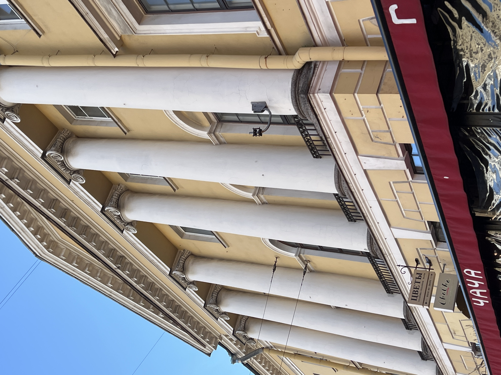 This is a low-angle, eye-level exterior shot of a building facade in Saint Petersburg, Russia. The building is painted a pale yellow and features a row of large, white, classical-style columns. Above the columns, there is an ornate cornice with dentil detailing, and a blue sky is visible in the upper left corner. Below the columns, a dark balcony railing runs horizontally. Further down, there are two signs hanging from the building. The upper sign is rectangular with text in Russian and appears to be for a flower shop. The lower sign is smaller and rectangular with text in a script font. A dark red awning extends from the building over what appears to be a storefront with reflective, crinkled material behind it. A white Cyrillic letter "Г" is visible on the awning. The overall impression is of a historic building in a commercial district.
