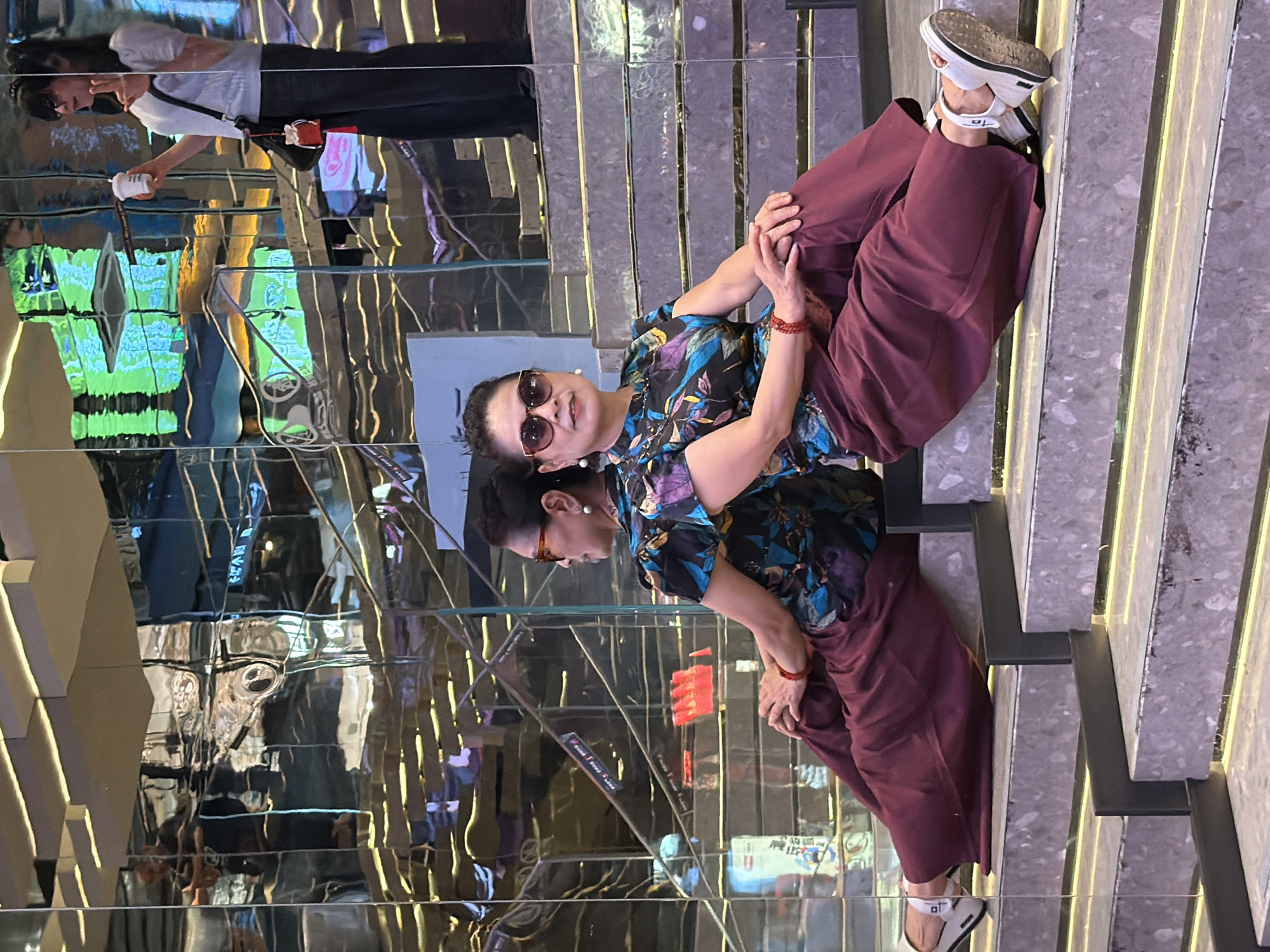 In Yuzhong District, China, two women are positioned on a set of modern stairs. The woman in the foreground, seated on the steps, faces the camera wearing sunglasses, a patterned top, and maroon pants. Her reflection is visible to her left, mirroring her pose. To the right and slightly behind her, another woman stands holding a white cup, also wearing sunglasses and a light-colored shirt. The environment is characterized by reflective surfaces, likely made of glass or polished metal, which create a distorted and multifaceted view of the surroundings, including stairs and lighting elements.