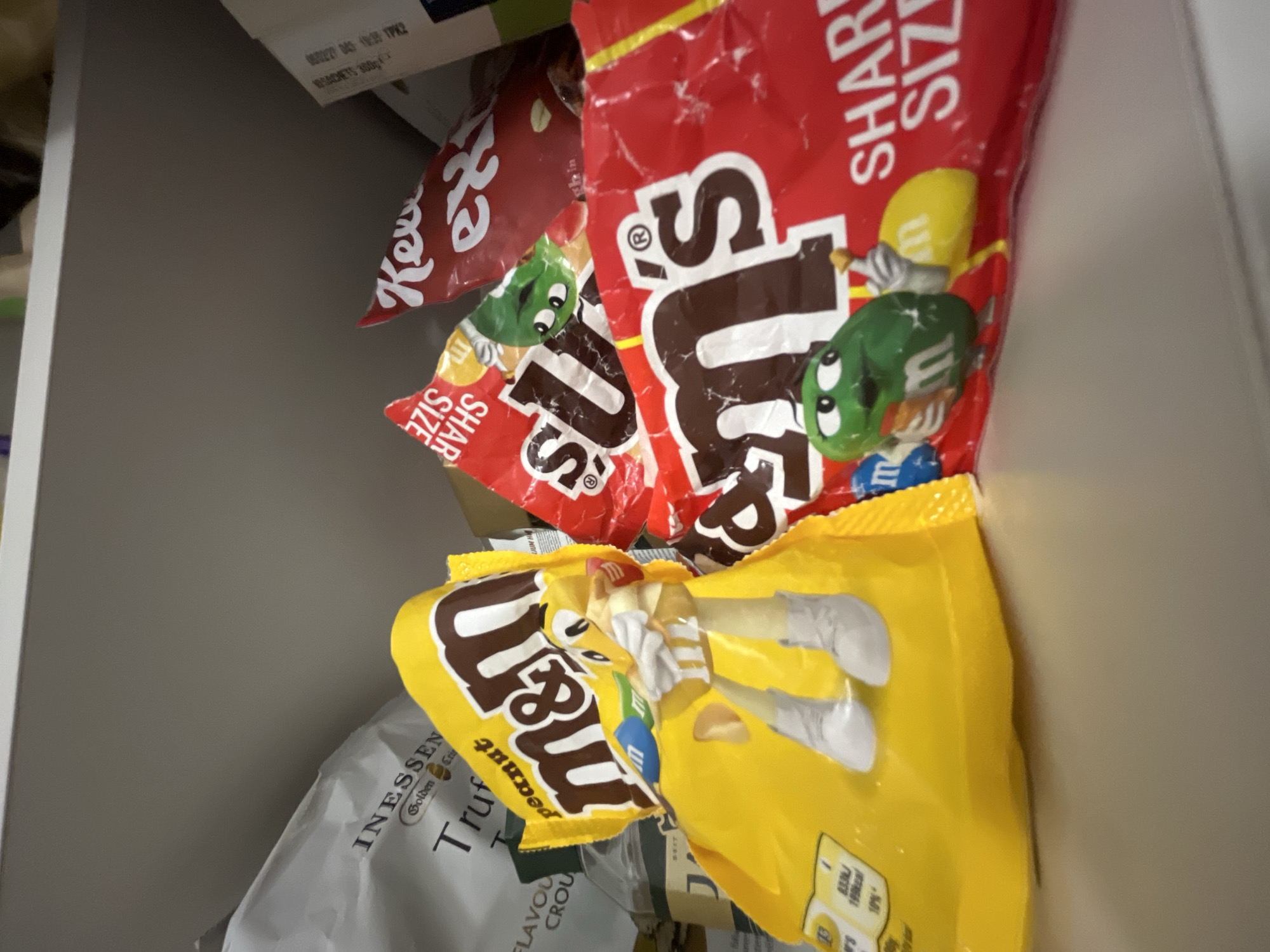 A close-up, low-angle, indoor shot shows several bags of M&M's candy on a shelf. The M&M's bags are primarily red and yellow, with one red bag featuring the M&M's logo and the words "SHARE SIZE" in white lettering. A yellow bag displays "M&M's peanut" and a cartoon M&M character wearing white boots. Another red M&M's bag, slightly out of focus, also shows the "SHARE SIZE" text. To the left, part of a white bag with text including "INESSEN" and "Truffles" is visible. The shelf is grey, and the lighting is soft, suggesting an indoor storage area, possibly a pantry or cupboard. The scene does not contain any information relevant to the city of Limassol, Cyprus.