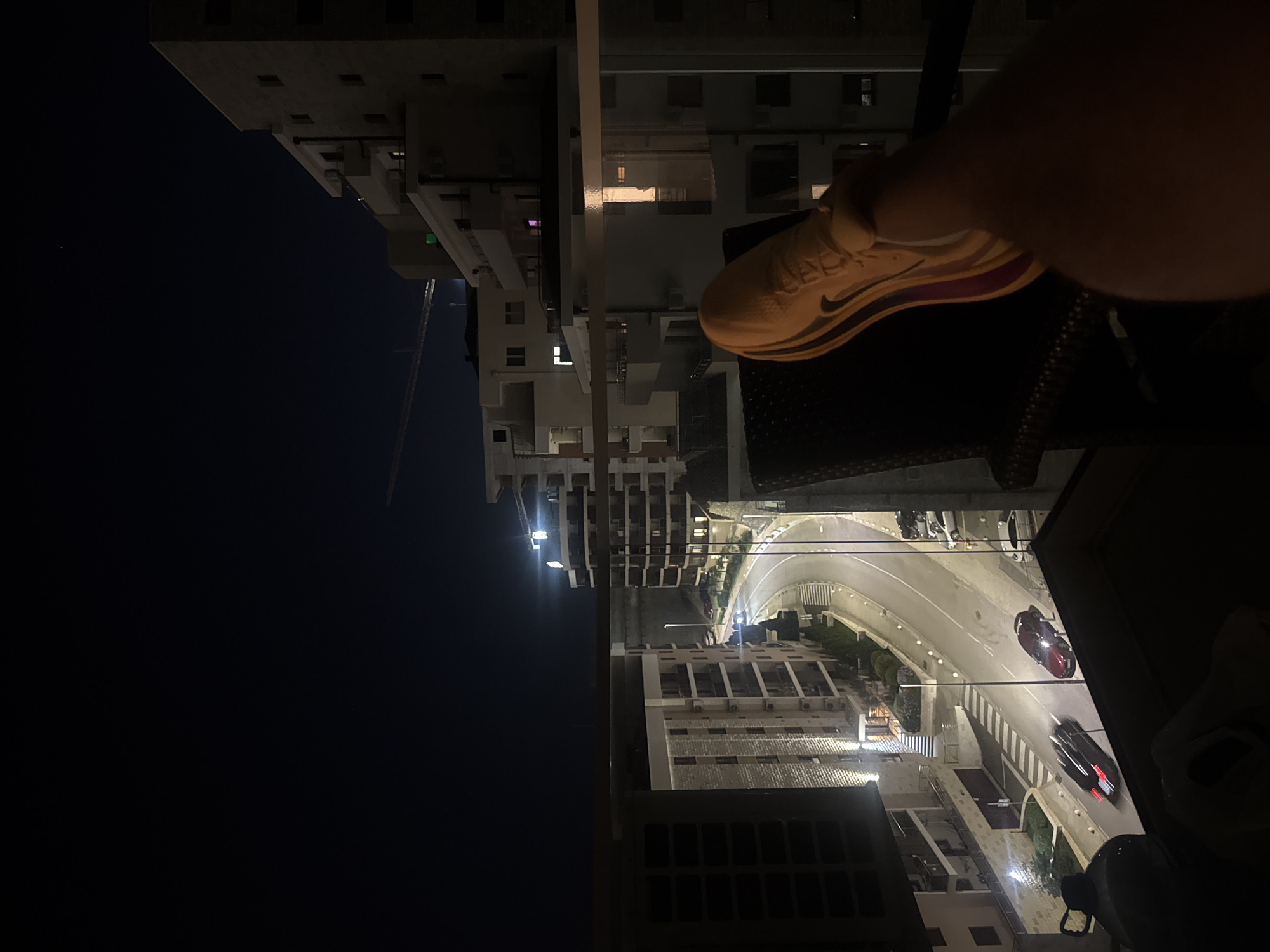 The image, taken at night from a balcony in Boreti, Montenegro, shows a view of a curving street lined with apartment buildings. A person's leg and foot, wearing a yellow athletic shoe, are in the foreground, resting on a chair. Cars are visible on the street, with one red car stopped and a black car in motion, its taillights blurred. The sky is dark, with a faint moon or bright light visible in the distance. A construction crane is also present in the background. The buildings have illuminated windows, suggesting habitation. The scene is illuminated by streetlights and the ambient light from the buildings.