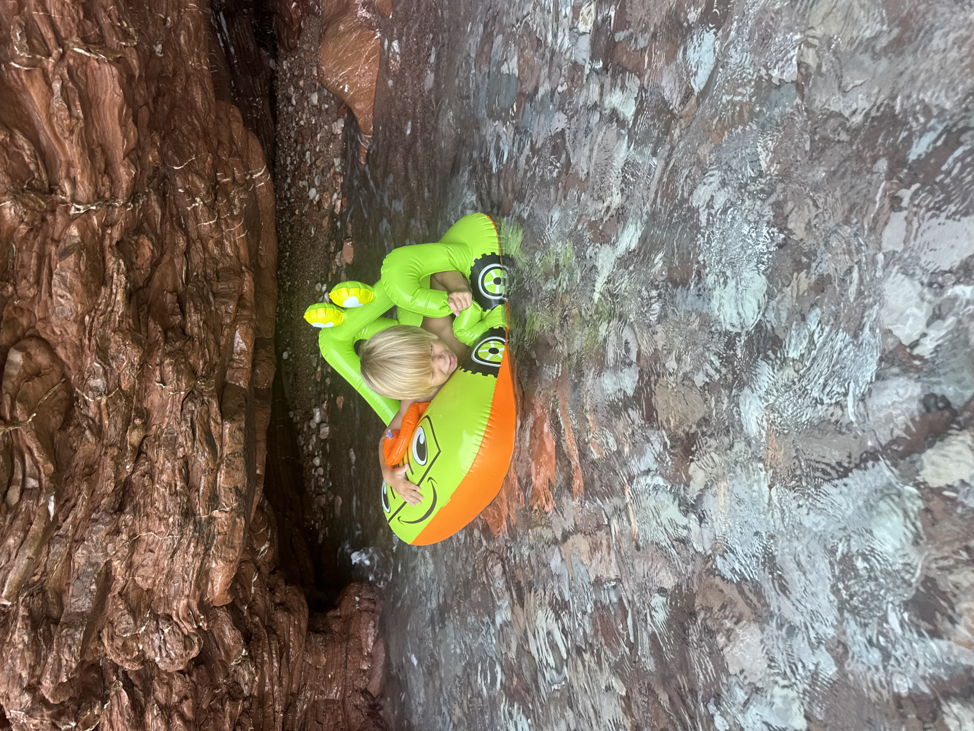 In this overhead shot, a young blonde child with fair skin enjoys a sunny day in the clear waters of Montenegro, near Sveti Stefan. The child is nestled within a vibrant green and orange inflatable toy shaped like a cartoon frog, complete with yellow eyes. The child is wearing a small orange safety vest. The setting appears to be a cove or a shallow bay, with textured, reddish-brown rocks on the left side of the frame and the rocky seabed visible beneath the transparent water. Sunlight glints off the water's surface, creating a shimmering effect and highlighting the clarity of the water, which reveals the submerged pebbles and rocks. The overall mood is one of peaceful recreation and enjoyment of nature. No visible text is present in the image.
