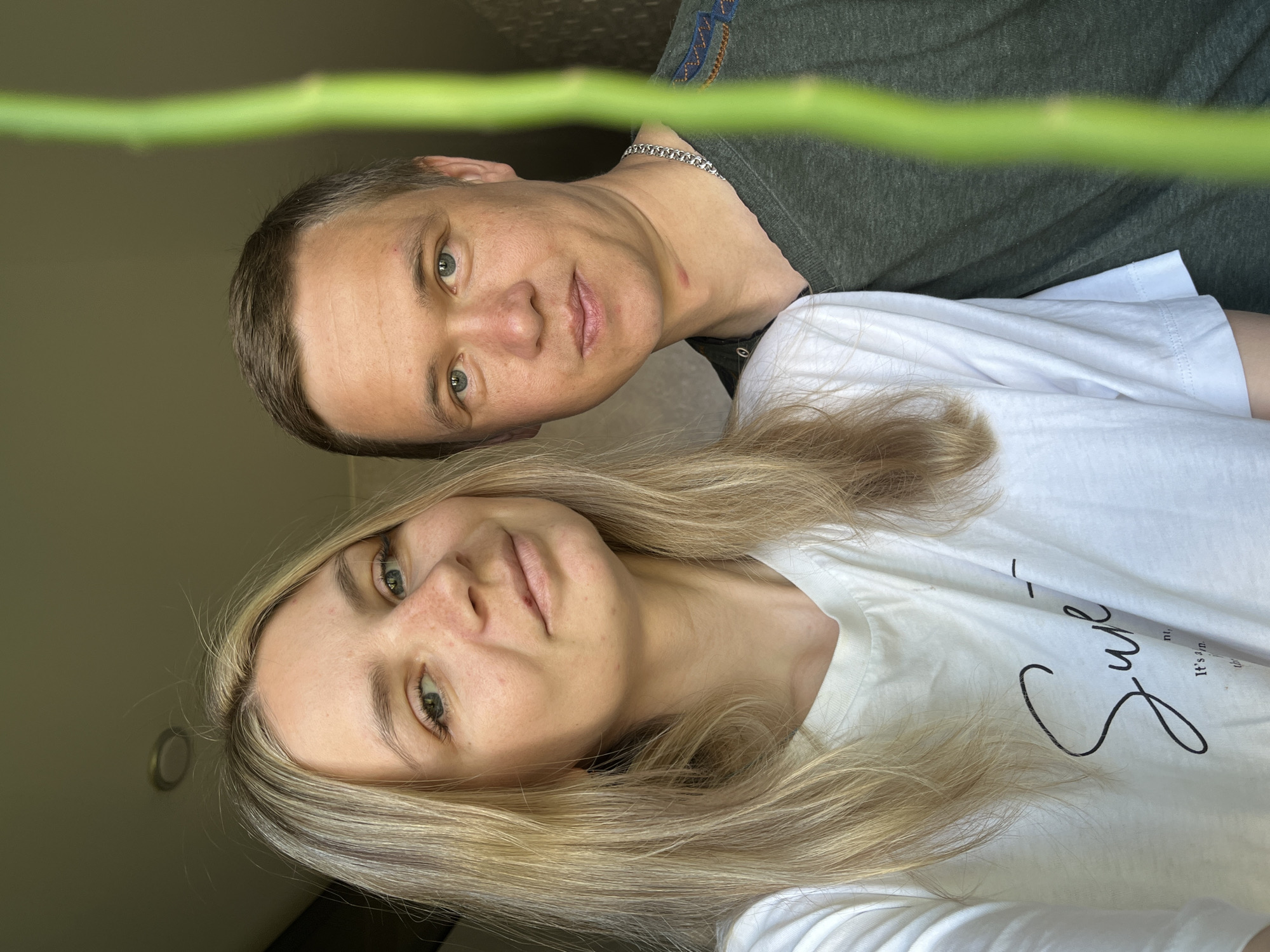 A selfie shot from a low angle depicts two individuals, a man and a woman. The woman is positioned in the foreground, with her blonde hair falling around her face and onto her white T-shirt, which features the word "Super" in a calligraphic script. The man is behind her, visible from the neck up, wearing a dark green T-shirt and a silver chain. Both subjects appear to be looking directly at the camera with a neutral expression. Between them, a green stem or branch is visible, seemingly obscuring the background. The setting appears to be indoors, with a glimpse of a light switch or outlet on the left side of the frame. The location is Chaikovsky, Russia.