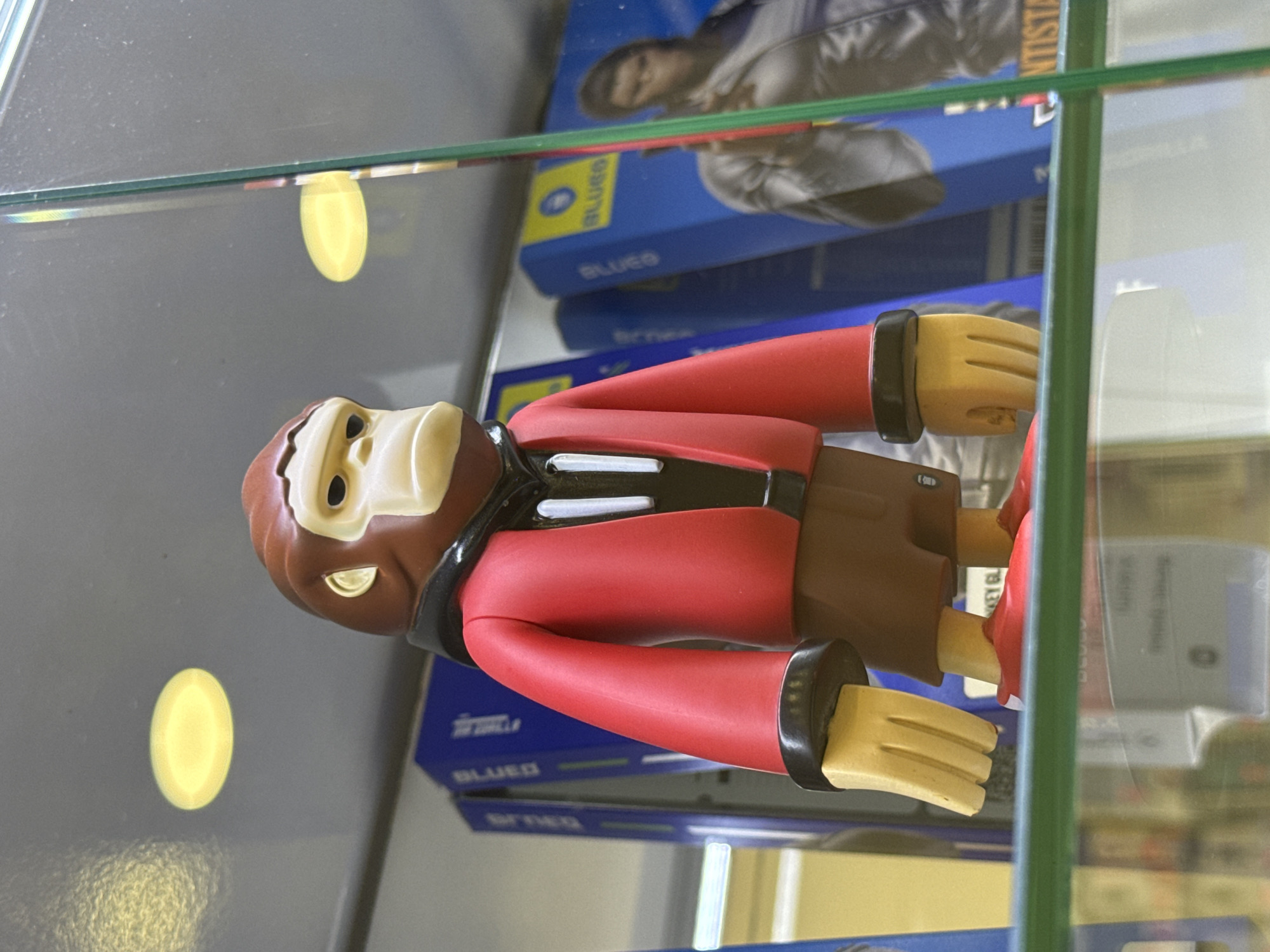 In Burgas, Bulgaria, an anthropomorphic monkey figurine with a brown head and white face is situated within a glass display case. The figurine is wearing a red jacket with black trim and brown pants. The hands are yellow. The background consists of blue and white boxes, some with text. There is a yellow circle reflection on the left side of the case.